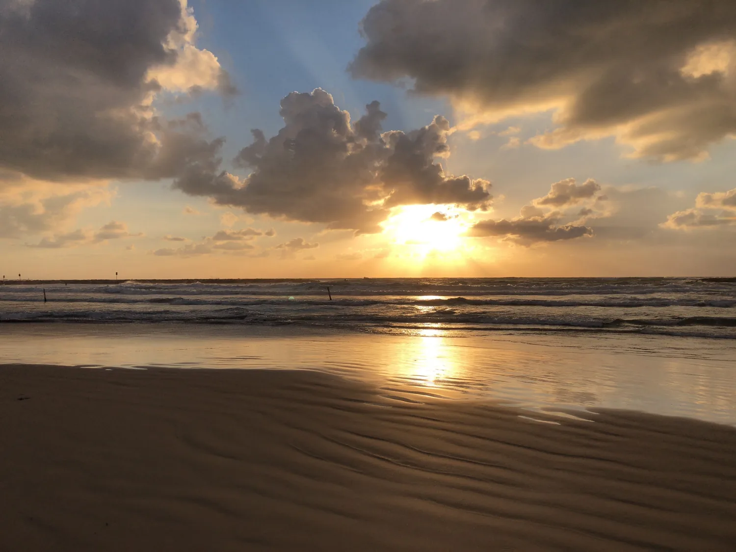 A beach at sunset seen low over the sand, the sun on the horizon, its light running toward the camera on the wet shore and cloud across the sky.
