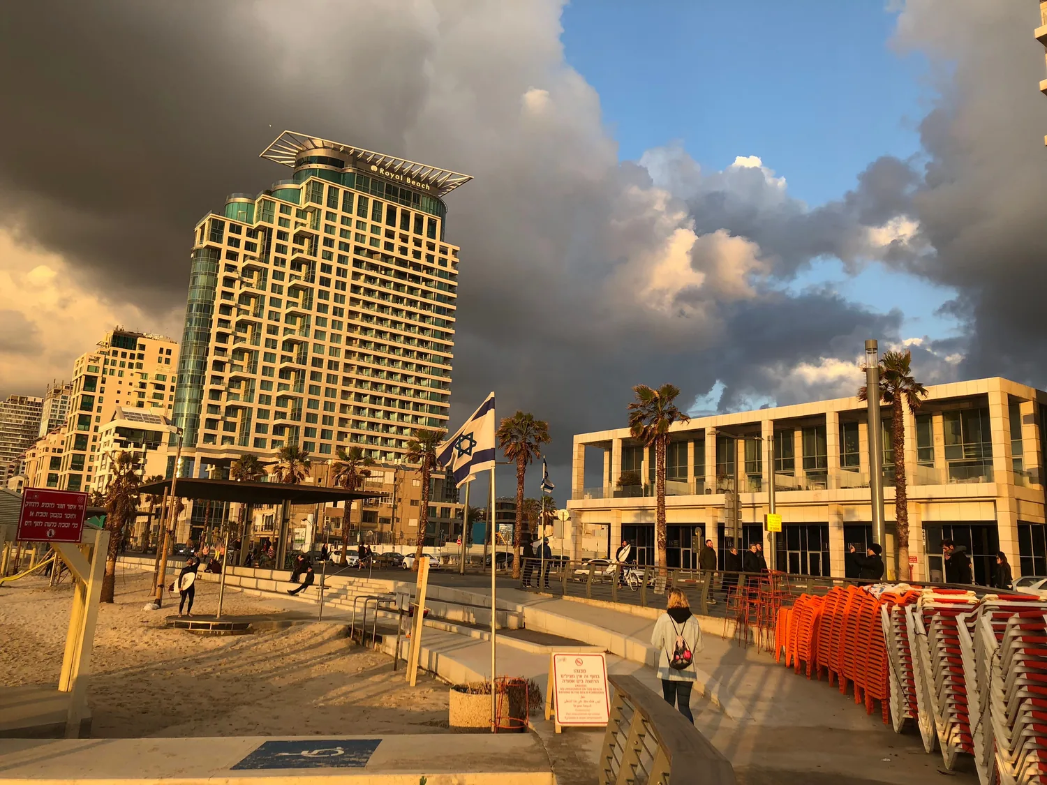 A seafront promenade in low golden light, tall buildings and palms along it, a flag on a pole and stacked chairs and tables on the paving.