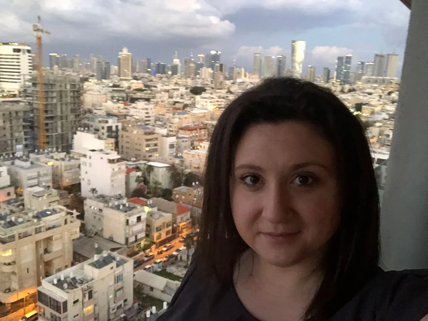A selfie of a woman with dark hair in a city high up, pale towers and rooftops packed together below her and more of the city out to the horizon.