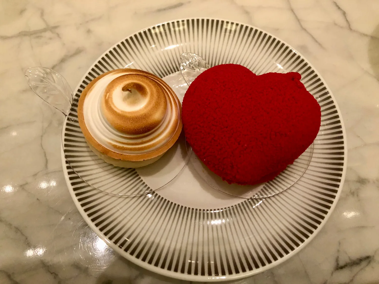 A white plate with a folded pastry on it and a small red cake beside it, on a marble table.