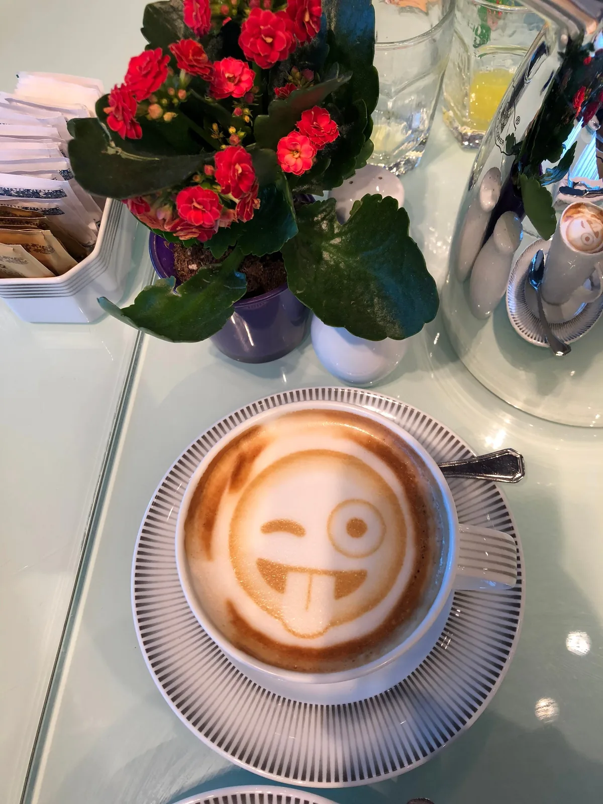 A cup of coffee on a saucer with a smiling face drawn in the foam, a small vase of red roses and a breakfast table behind it.