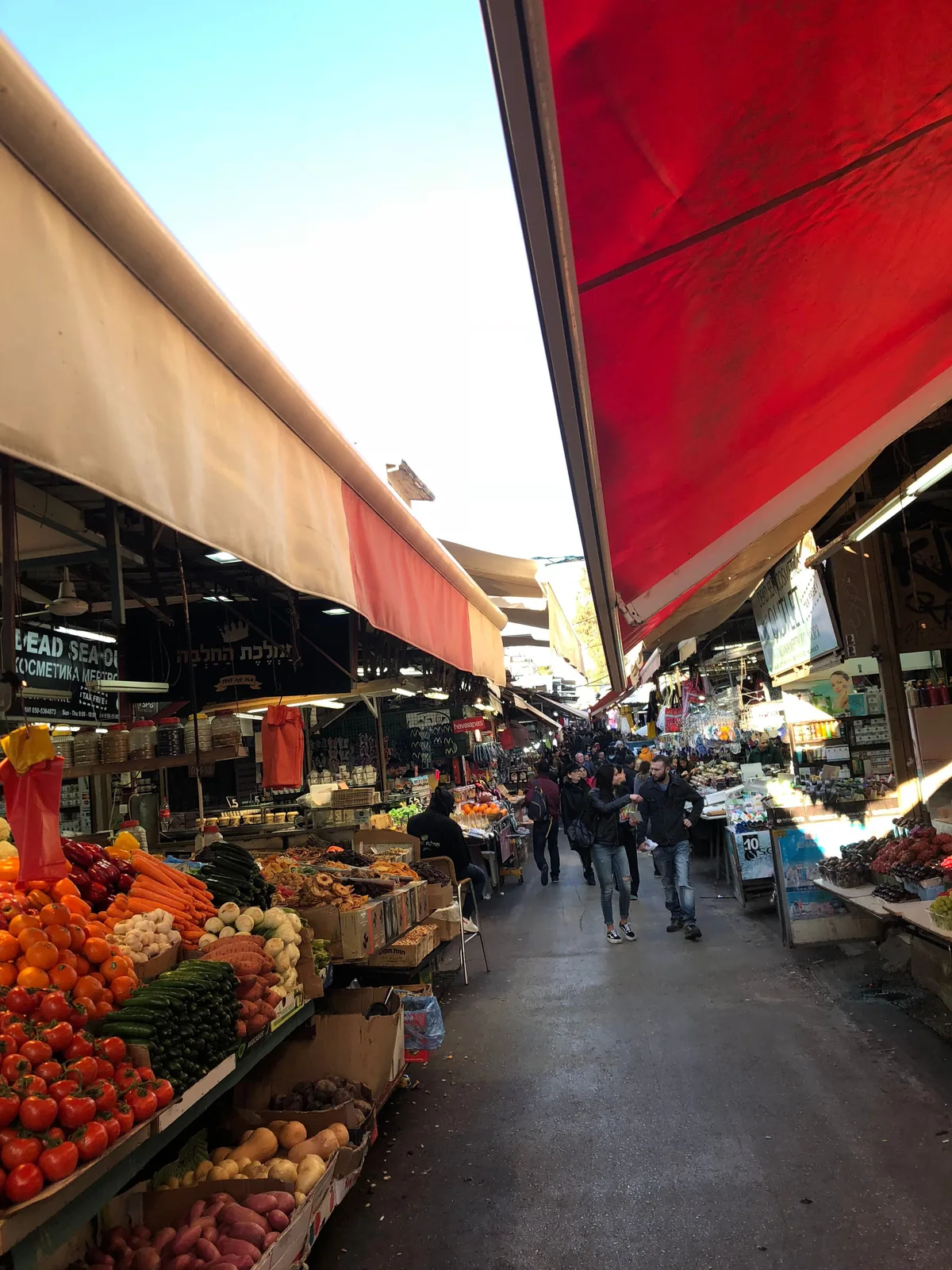 A market street under canvas awnings, produce stalls either side, shoppers walking away down the middle of the lane and a red awning overhead.