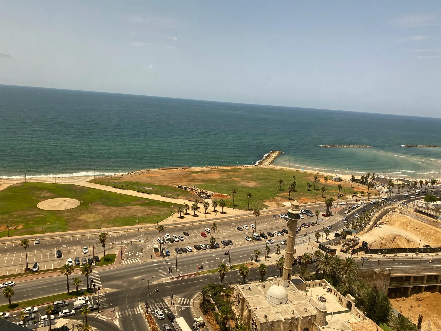 A seafront seen from high up, with a green lawn and a roundabout over road works below, a slim tower standing over the road and the beach and the sea beyond it.