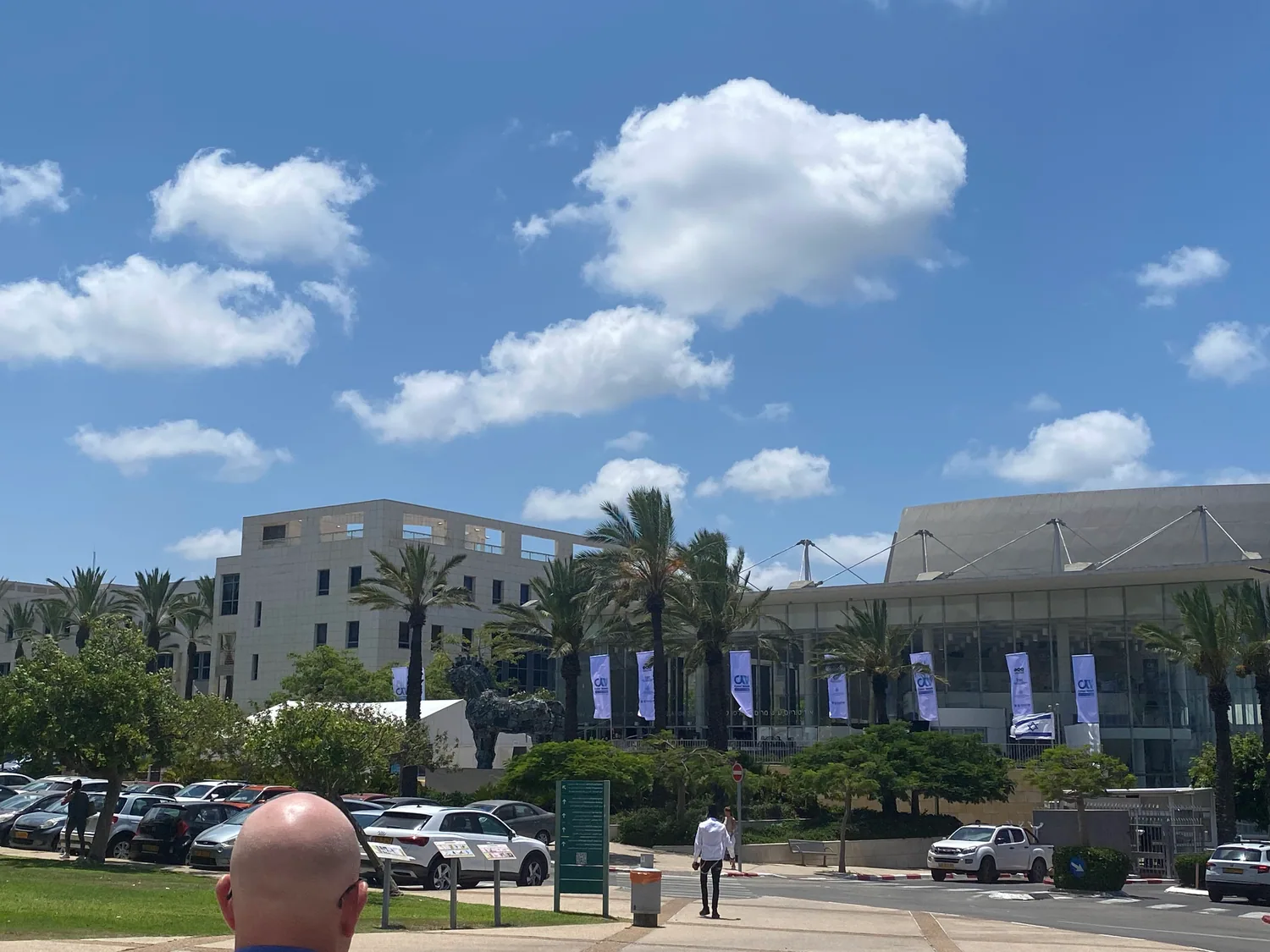A plaza between low buildings with palms along the front, cars parked at the kerb and stacked white cloud over the rooftops.