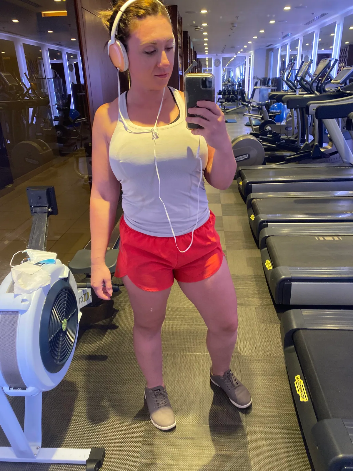 A mirror selfie of a woman in a white top and red shorts in a gym, headphones on and treadmills and machines behind her.