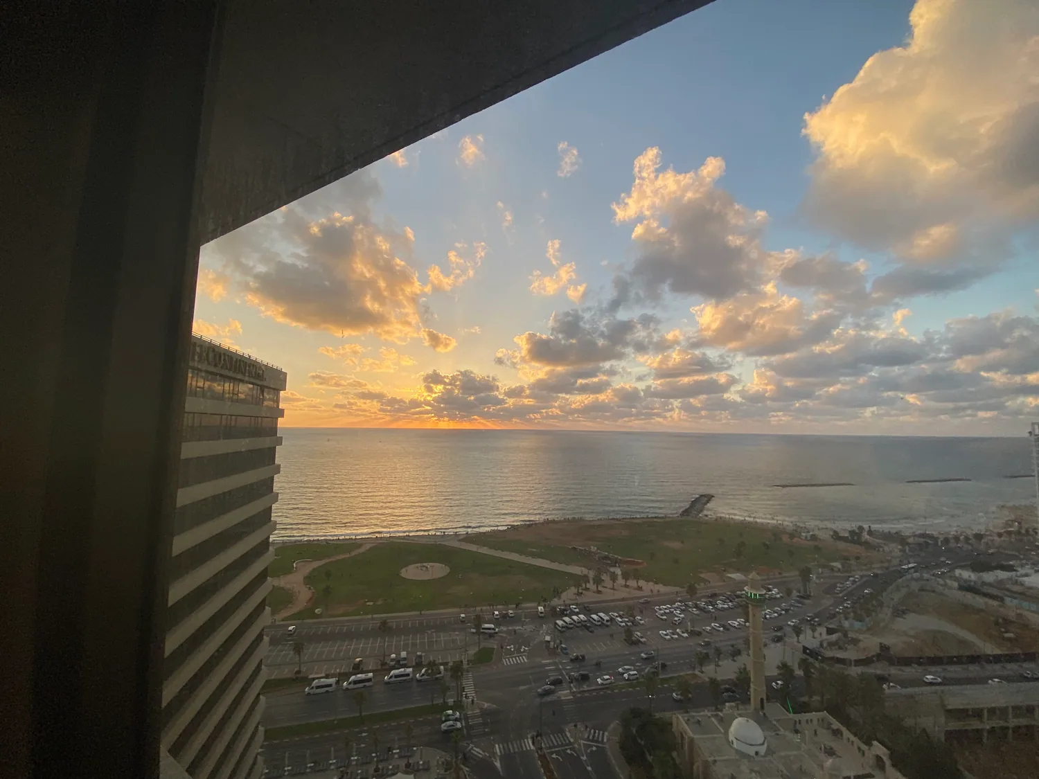 A bay and the sea at sunset seen from high up through a window, a town and its rooftops below and a wide band of cloud over the water.