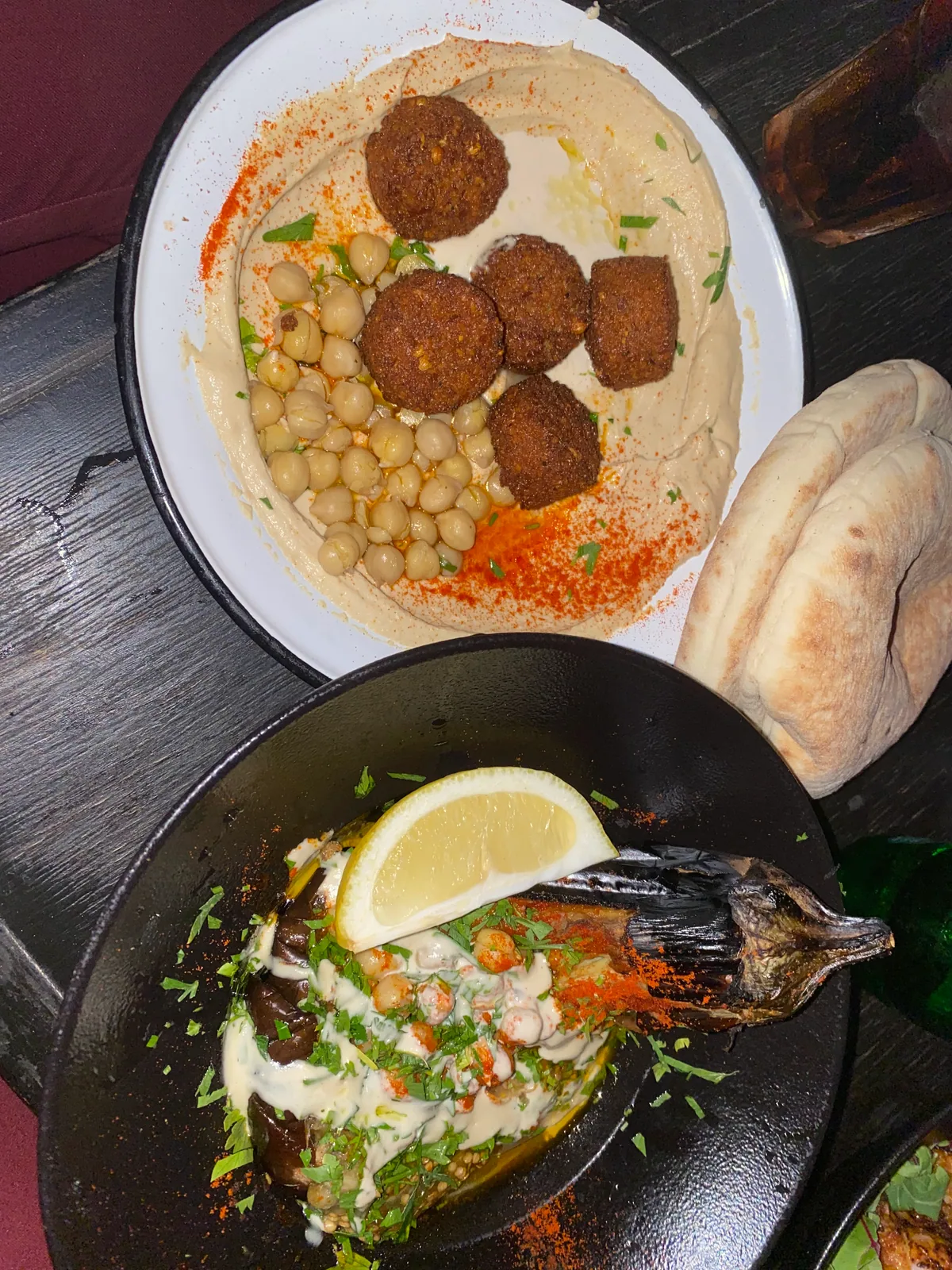 A plate of whole fish with lemon and herbs next to a plate of hummus topped with falafel, and bread at the edge of the table.