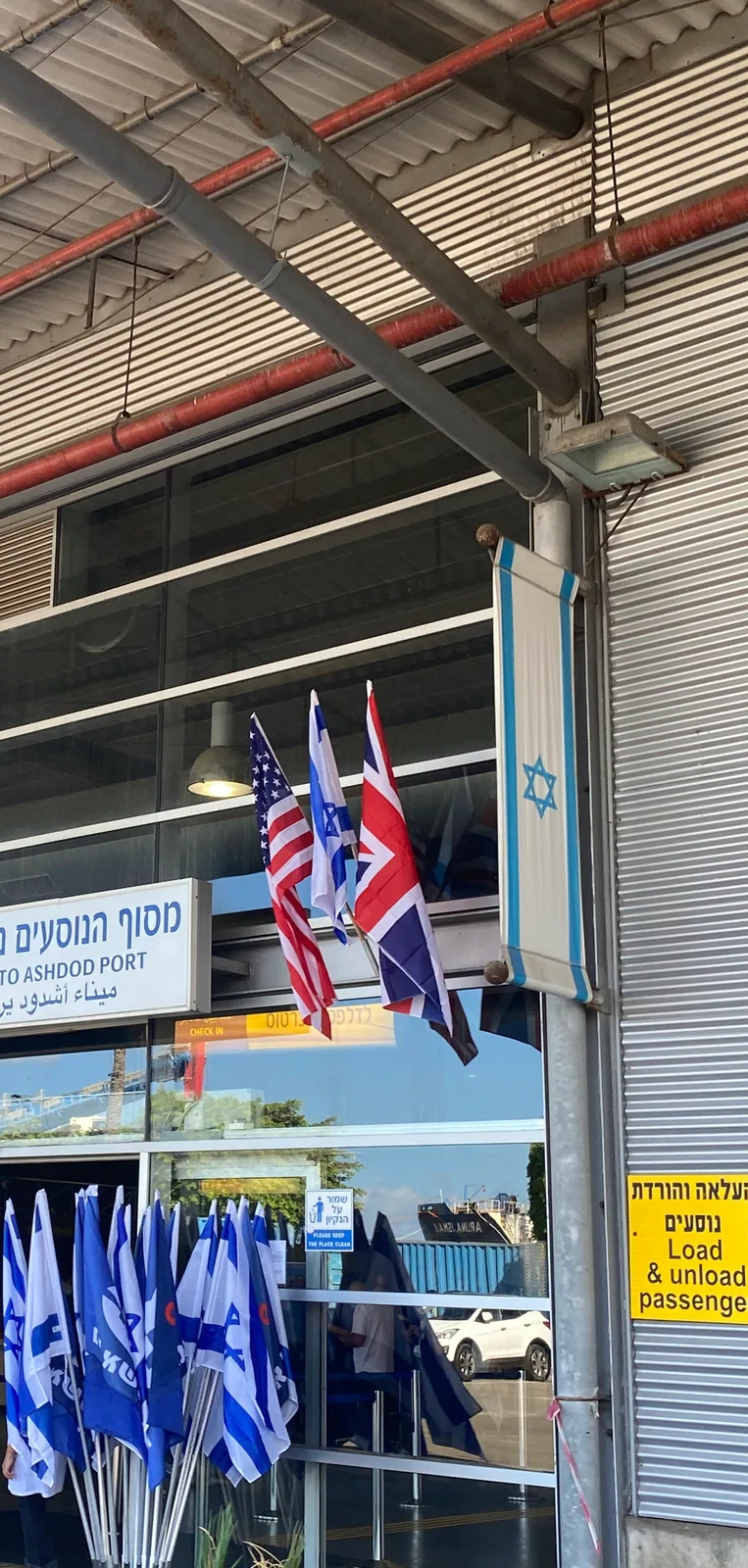 The entrance of a port with its welcome sign in three languages over the doors, flags of several nations on poles beside it and a check-in sign over the glass.