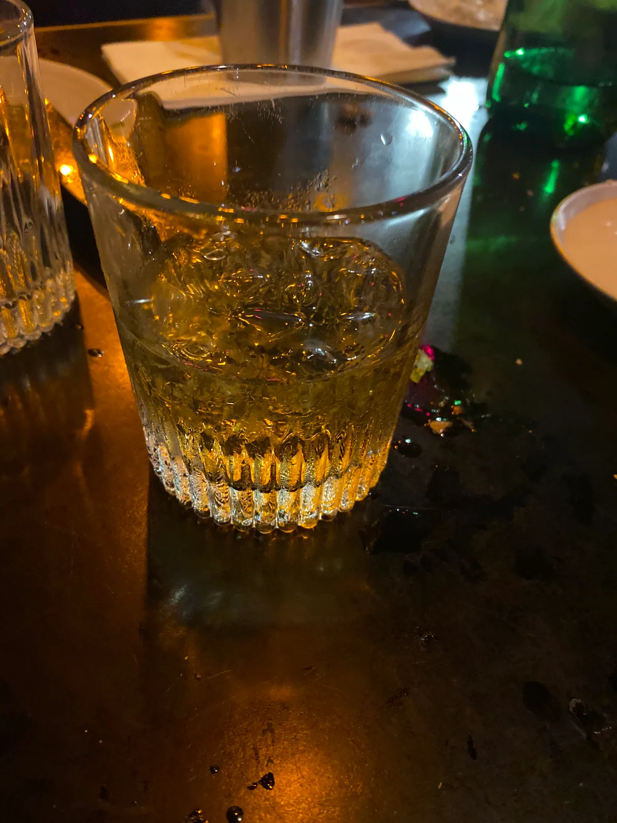 A glass of amber spirit on a dark polished table with a second glass at the left and bottles against the wall behind.