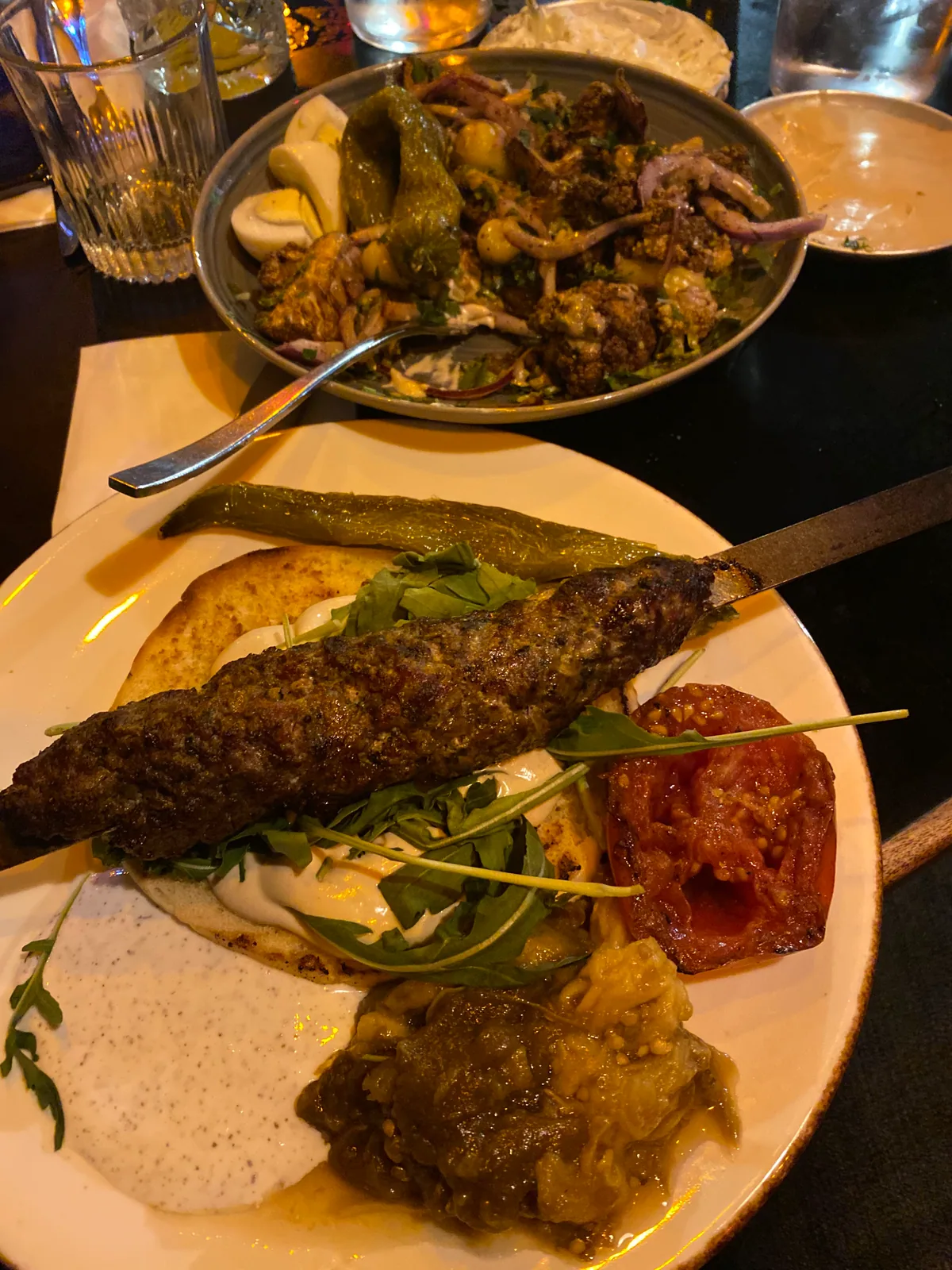 A plate of kebab with aubergine, tomato and bread on a table, with more dishes and a glass at the edge of the frame.