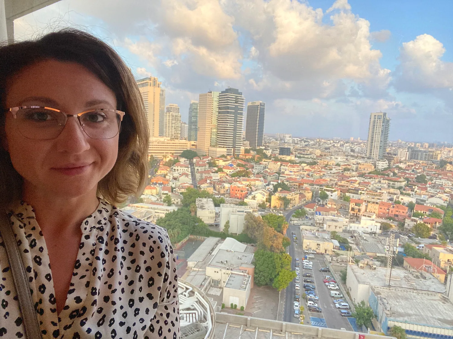 A selfie of a woman in glasses and a spotted blouse in front of a window, city rooftops and towers spread out behind her.