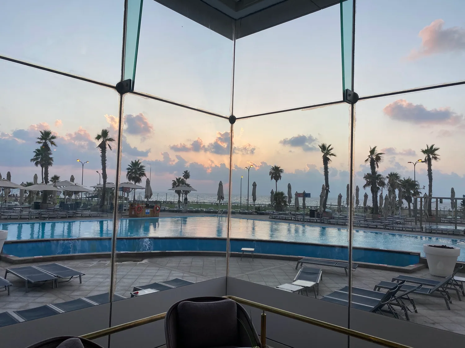 A pool deck seen through a glass wall at dusk, empty loungers along the water, palms along the edge and the sea beyond them.