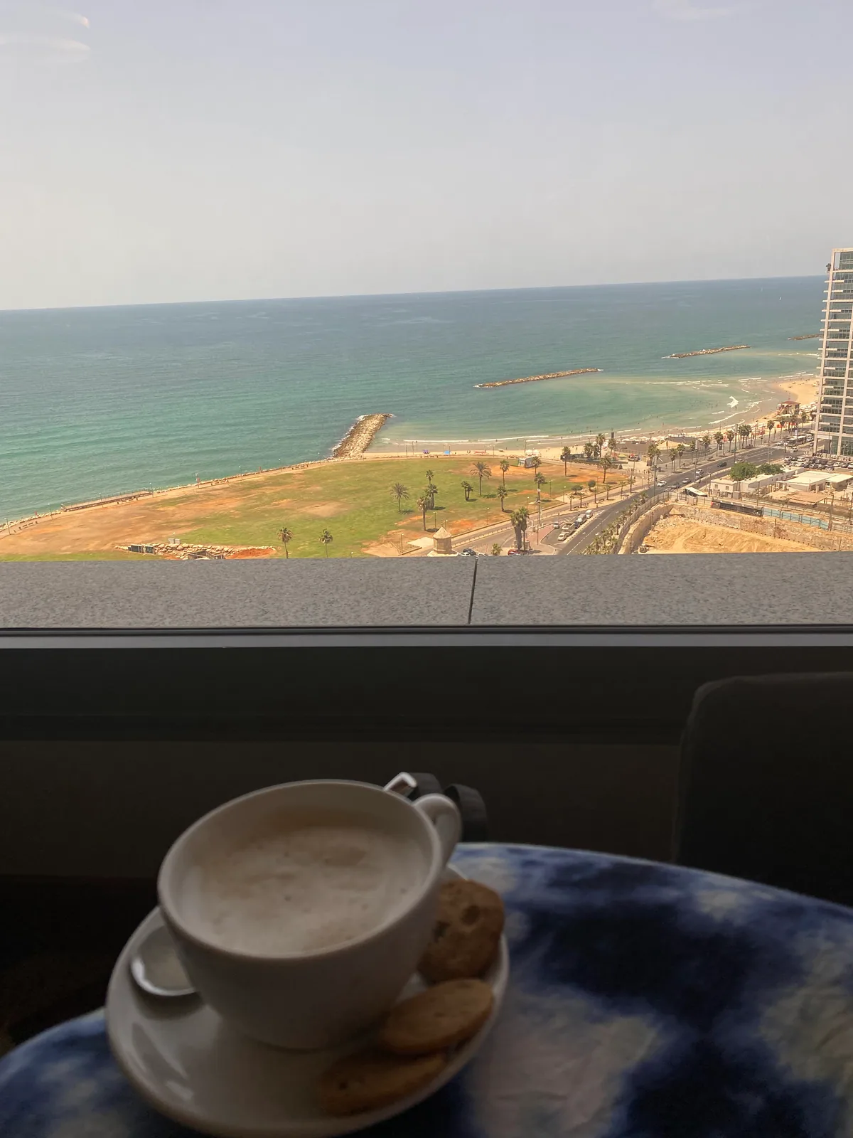 A cup of coffee and two biscuits on a blue patterned table beside a window, the sea and a breakwater out beyond the glass.