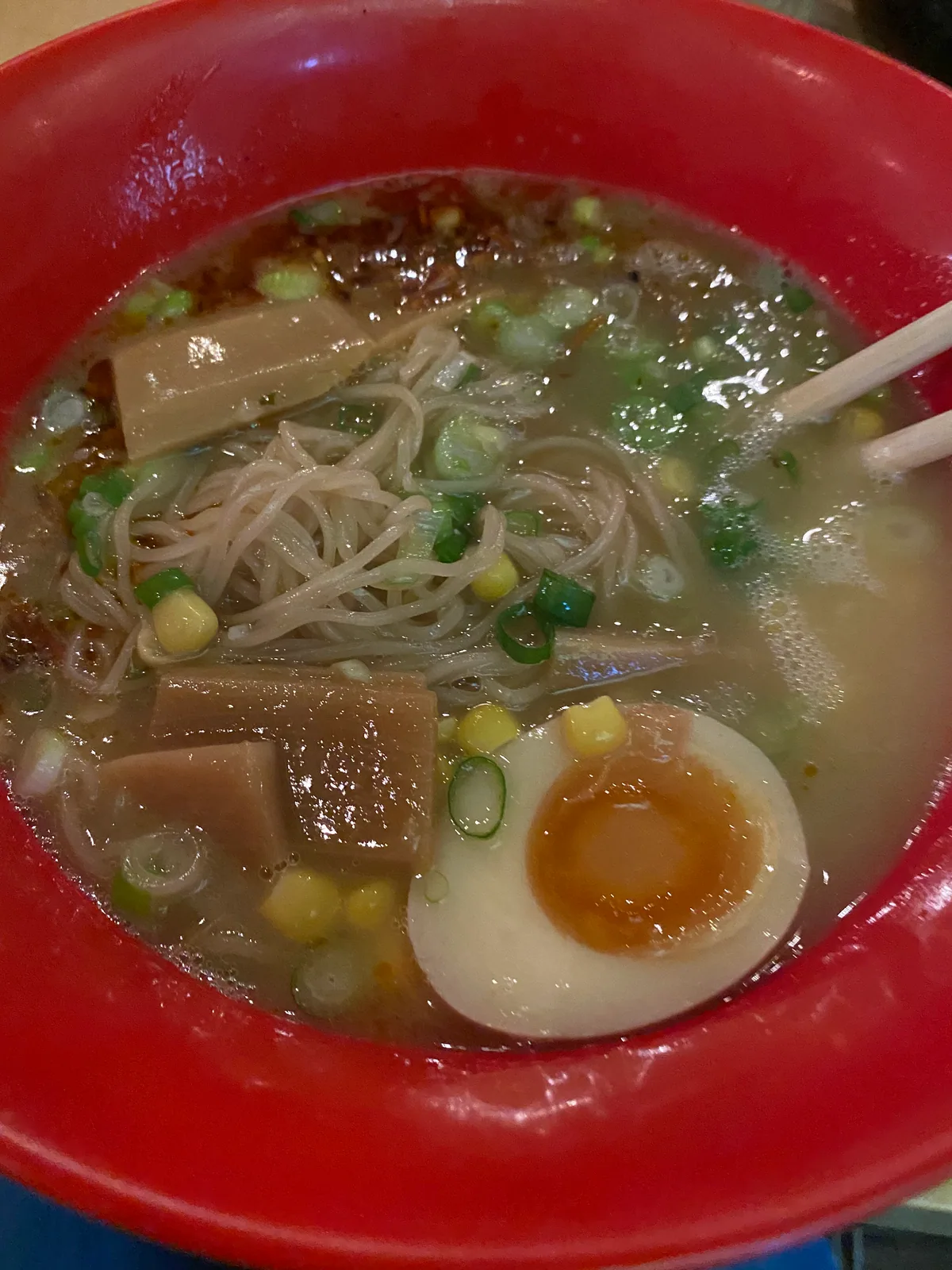 A red bowl of noodles with a soft-boiled egg, greens and broth, seen close up on a table.