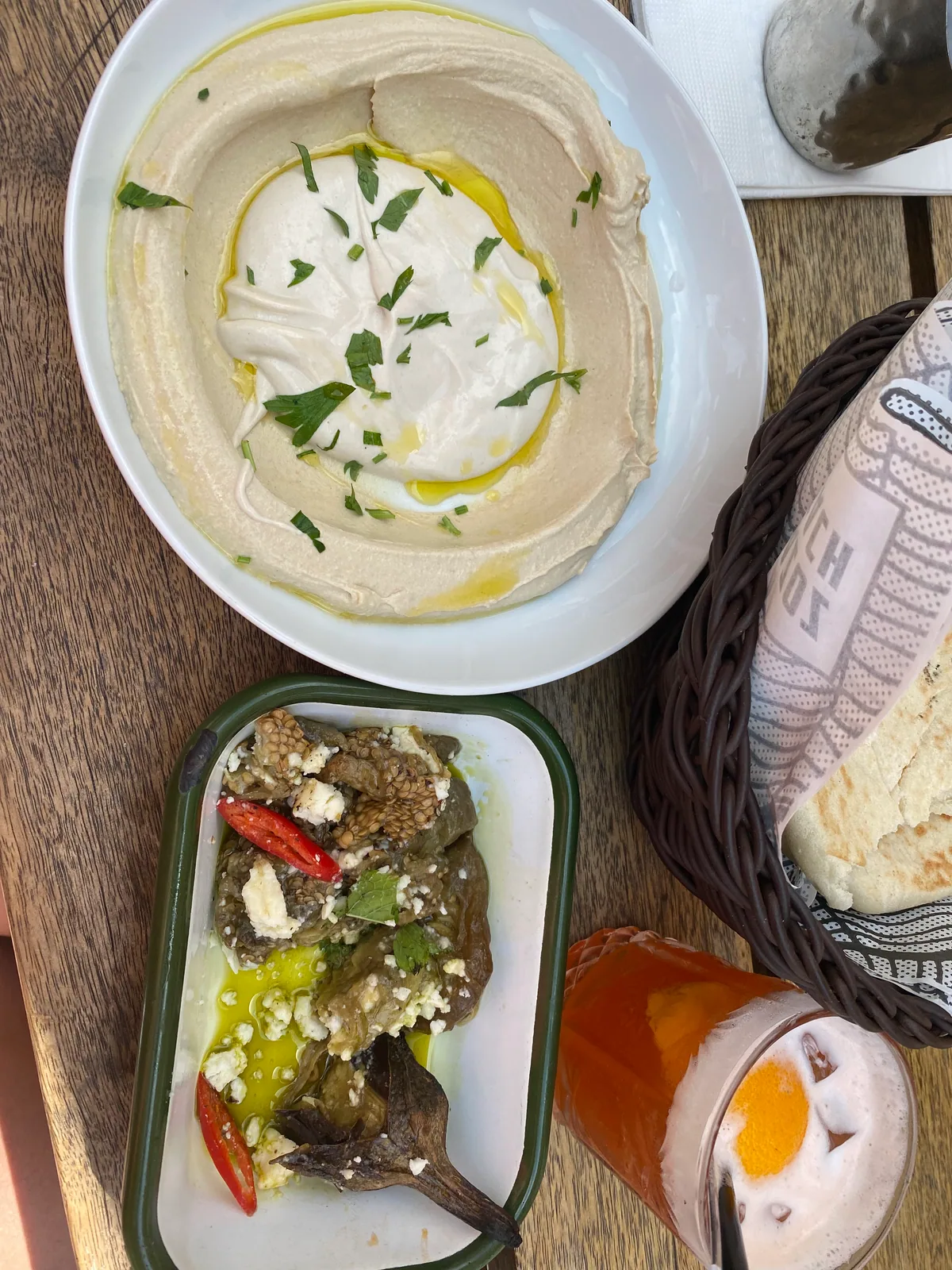 A bowl of hummus and a small dish of aubergine with bread coming out of a printed paper bag on a table, an iced drink beside them.