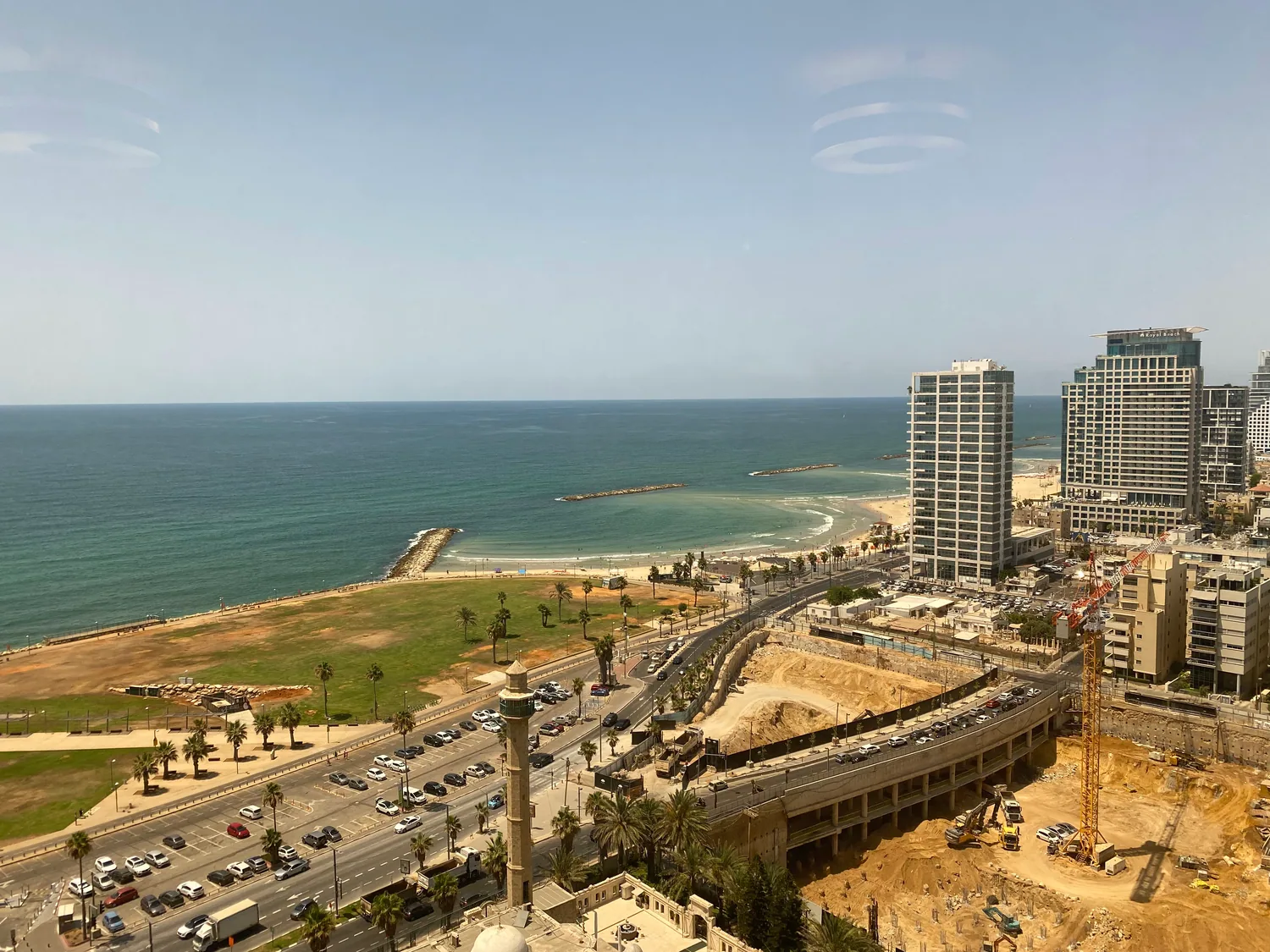 A coast seen along from a window, towers and a marina over road works below, and a breakwater running out into the sea.