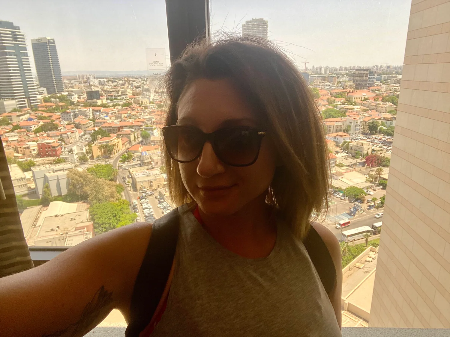 A selfie of a woman with dark glasses and dark hair in front of a tall window, city rooftops and towers behind her and a reflection in the glass.