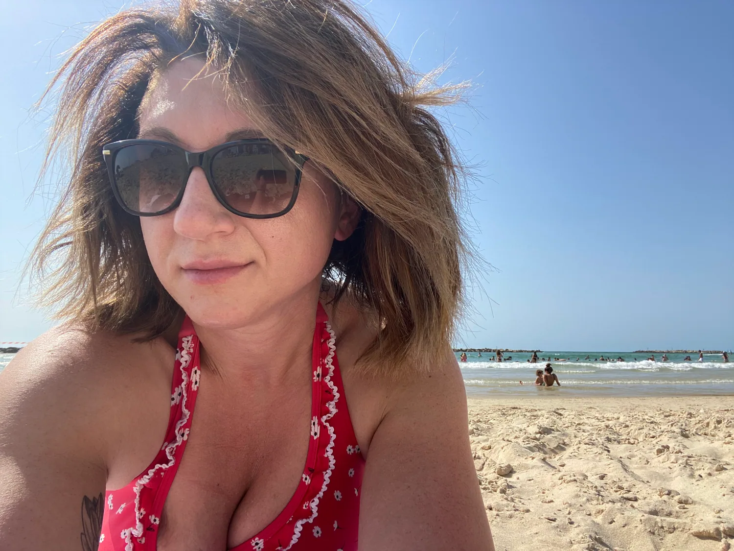 A selfie of a woman in a red patterned top and dark glasses on a beach, the sea and two people far behind her in the water.