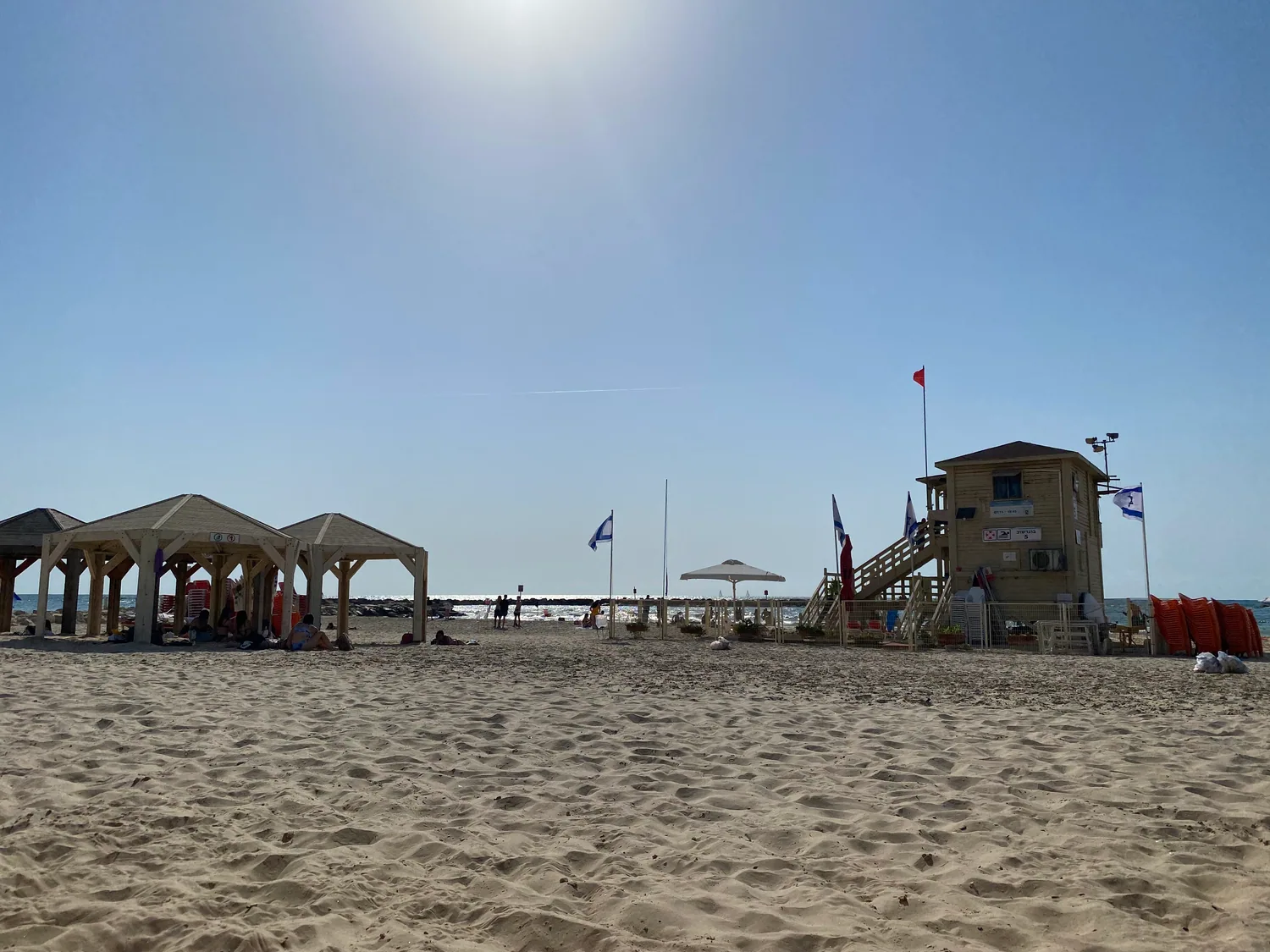 Three wooden pergolas standing in a row on the sand, small signs fixed to their posts, a tower behind them and the sea along the shore.