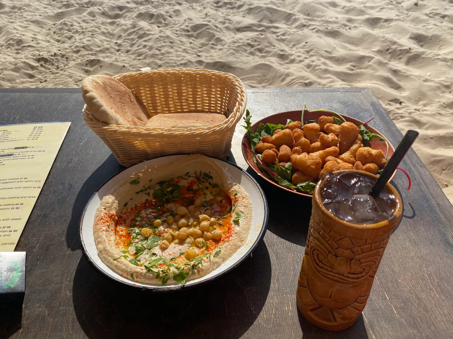 A table at the edge of the sand with a bowl of hummus, a plate of falafel, a basket of bread and a tall iced drink on it.
