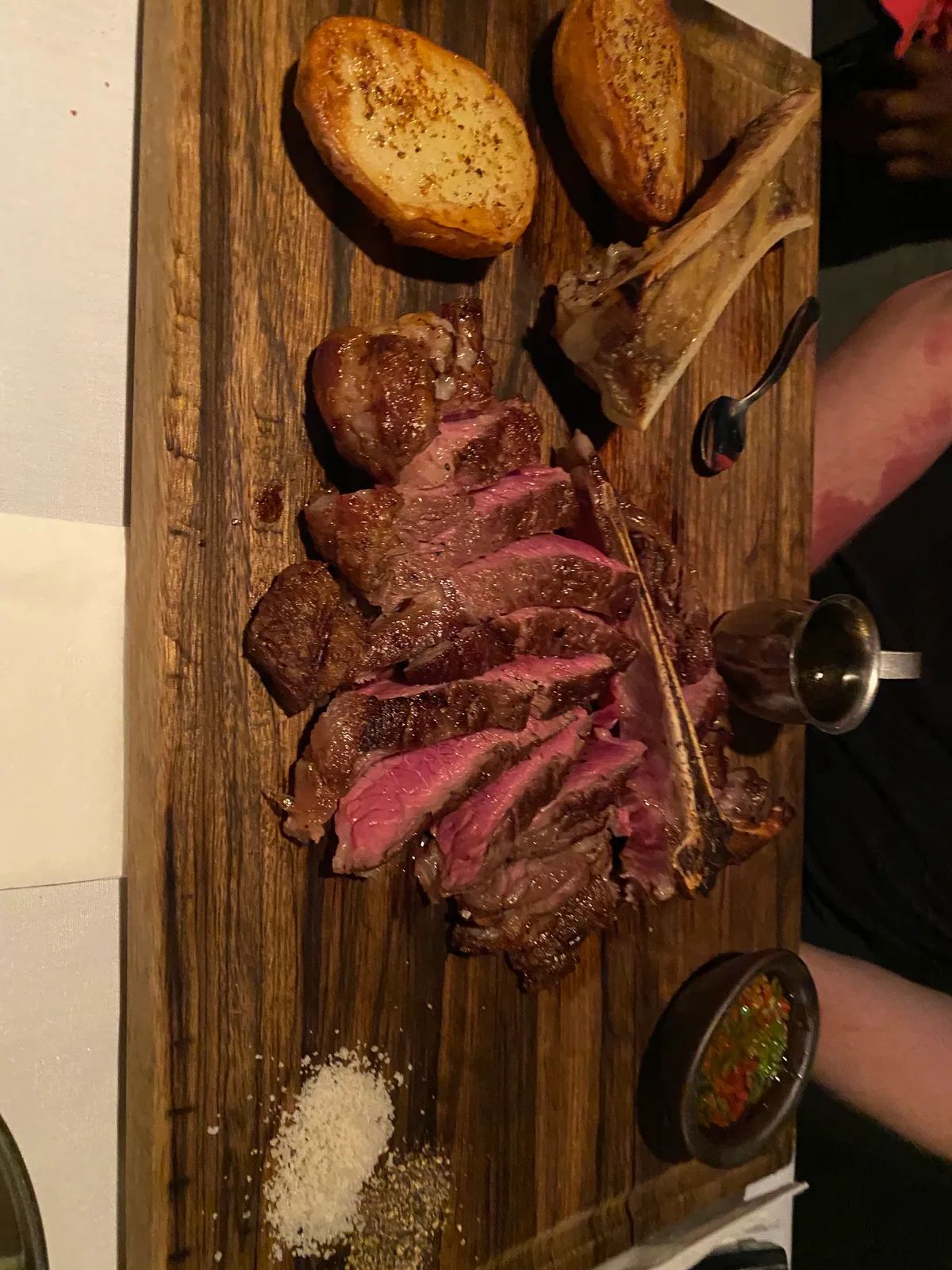 A sliced steak on a wooden board with bread, salt, a glass of wine and a hand holding cutlery at the edge of the frame.