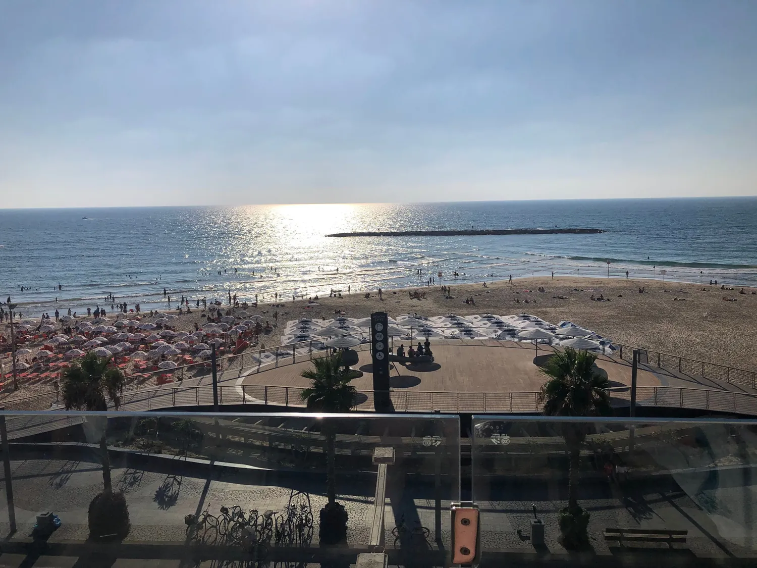 A beach and a promenade seen from high up, the sun on the water and its light running in toward the shore, rows of umbrellas on the sand, a round deck below and a glass balustrade across the front.