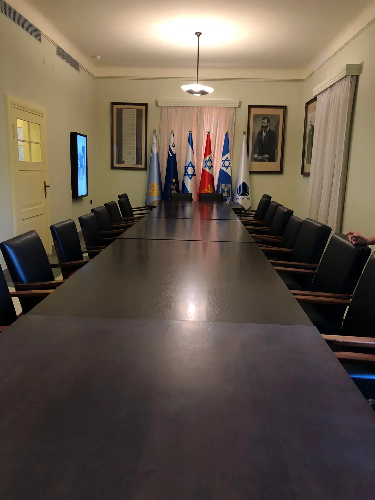 A long dark table running away down a room with chairs set along both sides, a curtain at the door, framed pictures on the walls and a rank of flags standing small at the far end.
