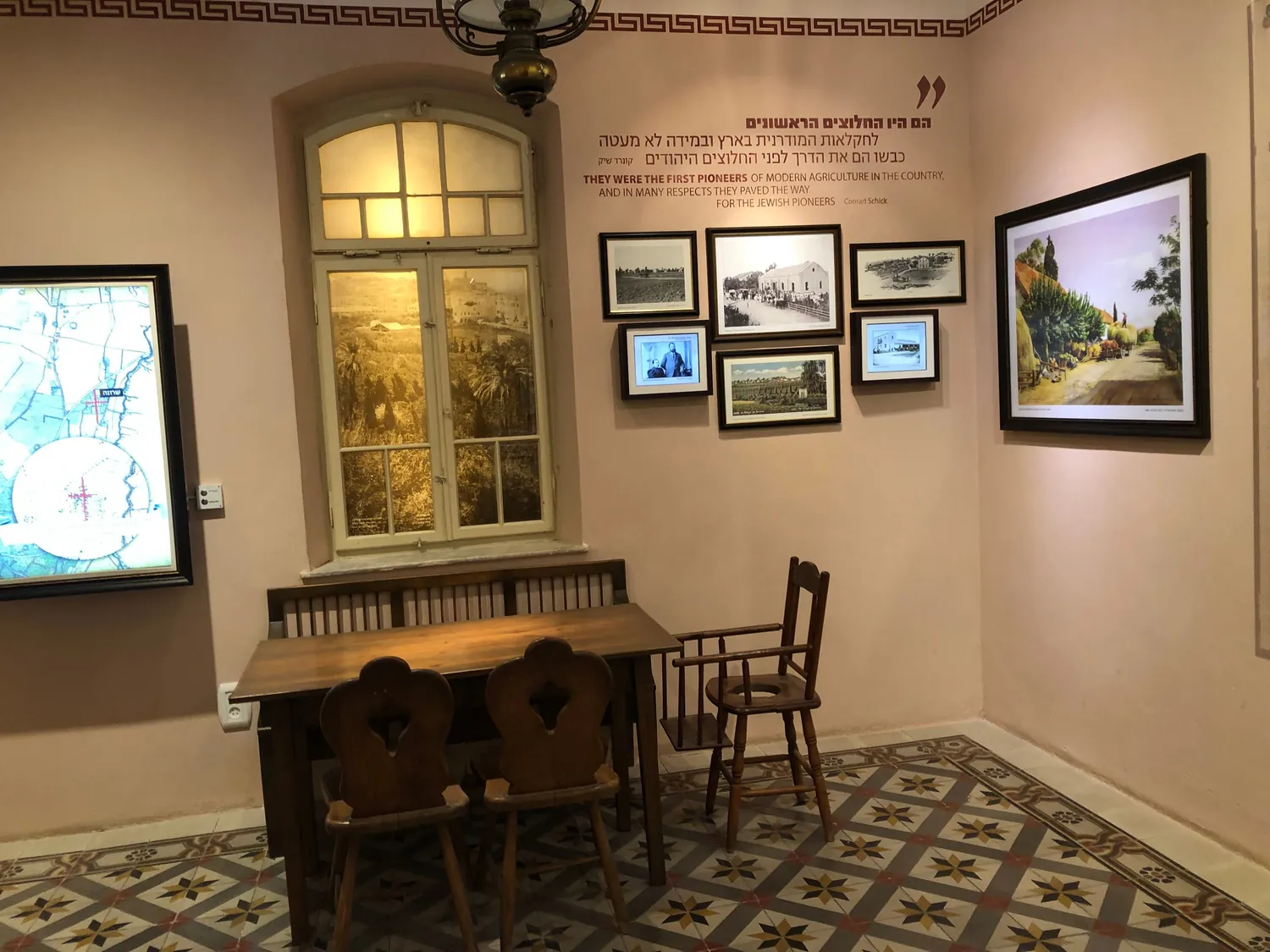 A room with a tiled floor and a patterned rug, a desk and two chairs, an arched window at one side and the walls hung with framed pictures and lettering.