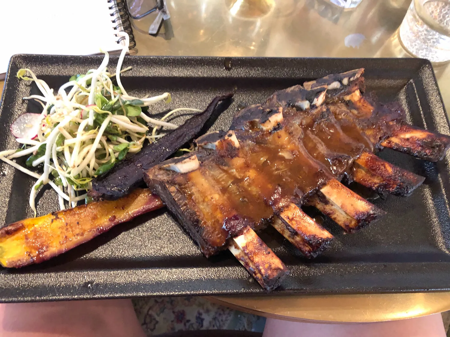 A board of glazed ribs with dressed slaw on a table, a hand holding the board at the bottom edge and a glass and cutlery behind it.