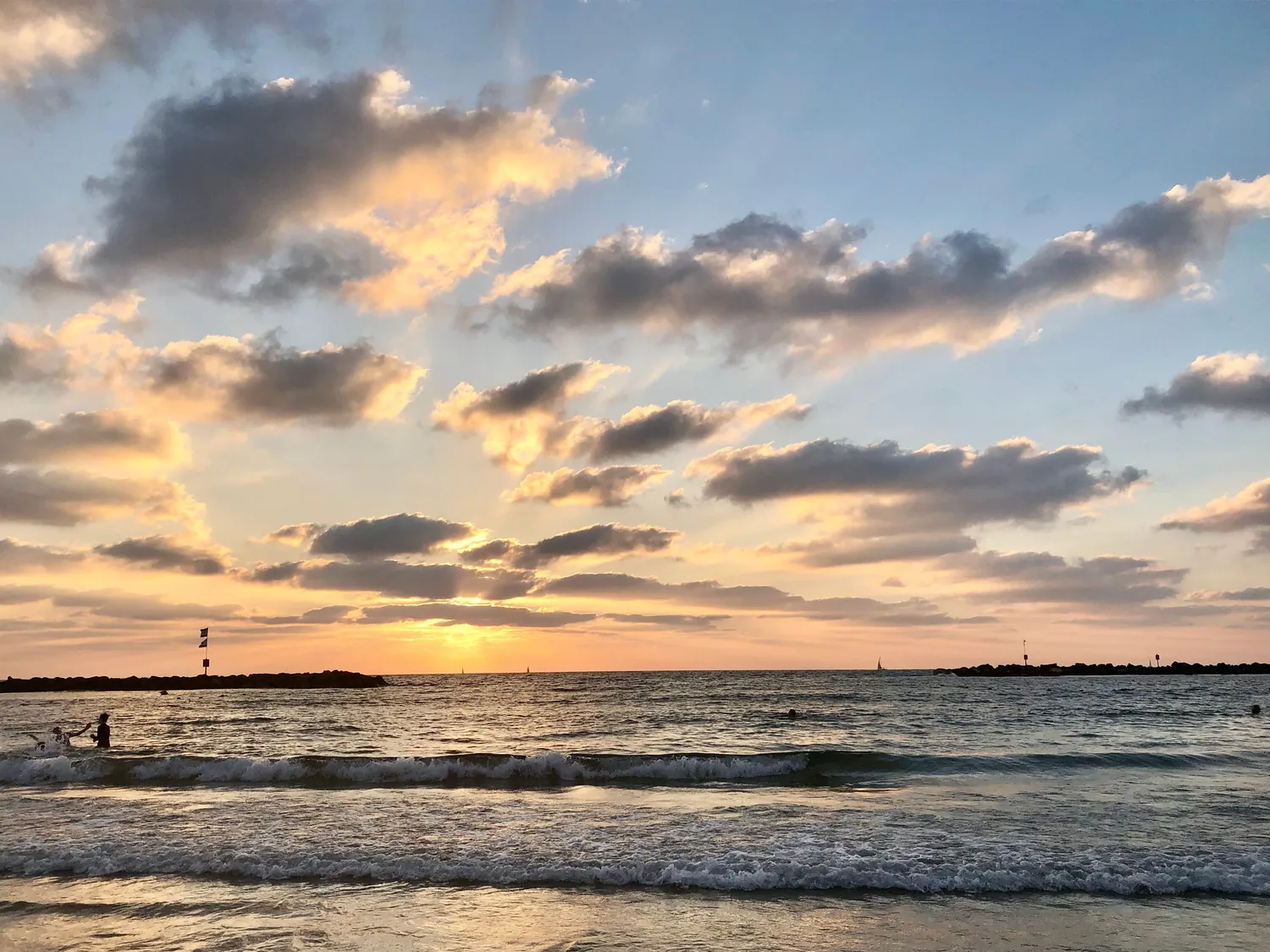 The sun going down over the sea under a wide bank of cloud lit gold, its light on the water and a breakwater running out from the shore.