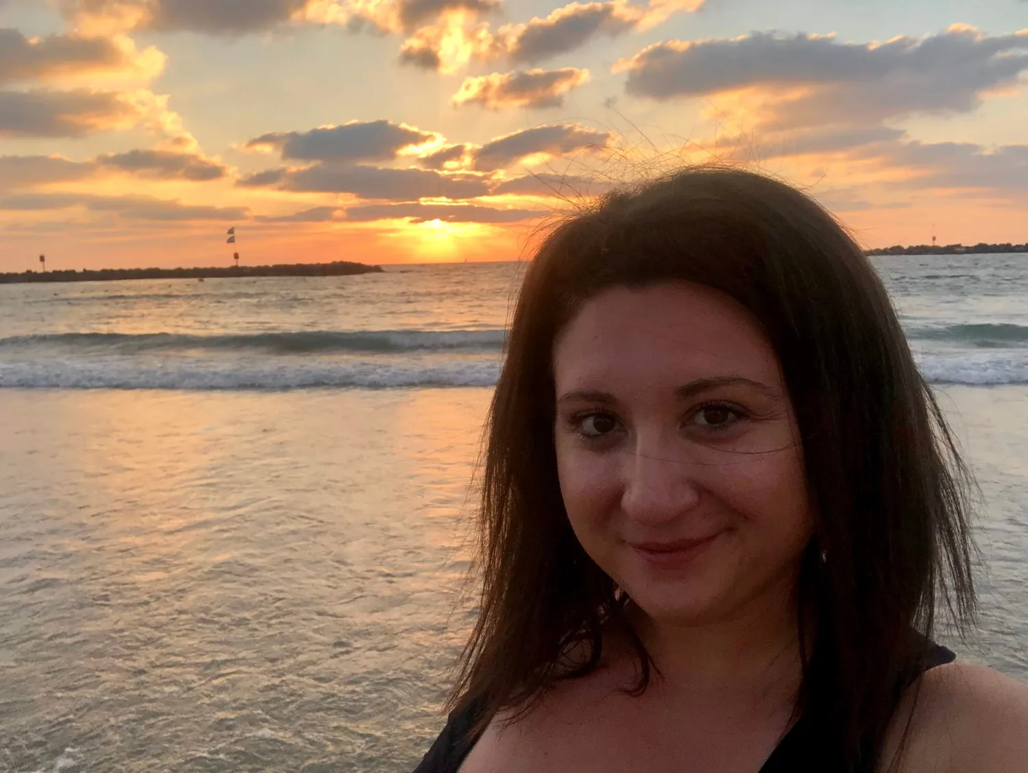 A selfie of a woman with dark hair on a beach at sunset, the sea and a breakwater behind her and cloud lit up across the sky.
