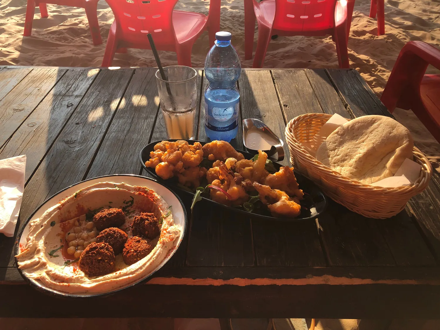A wooden table with a plate of falafel, a basket of bread, chips and a bottle of water on it, and empty red plastic chairs behind.