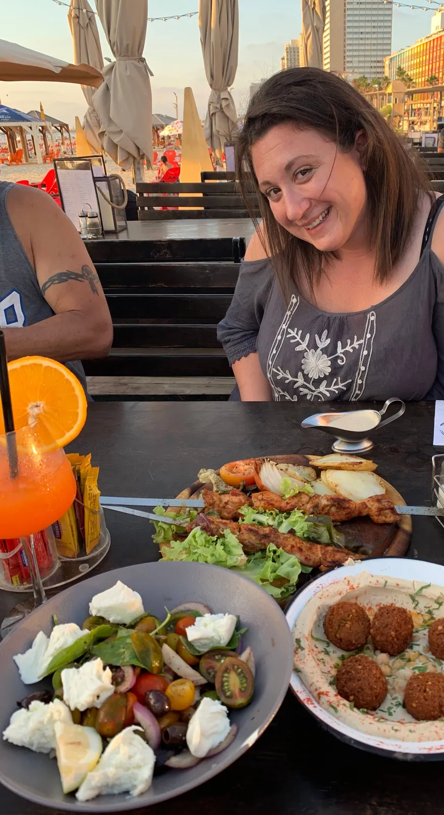 A woman with dark hair smiling at a table at dusk, plates of salad, falafel and skewers in front of her, a glass of orange juice at her elbow and a lit terrace behind.