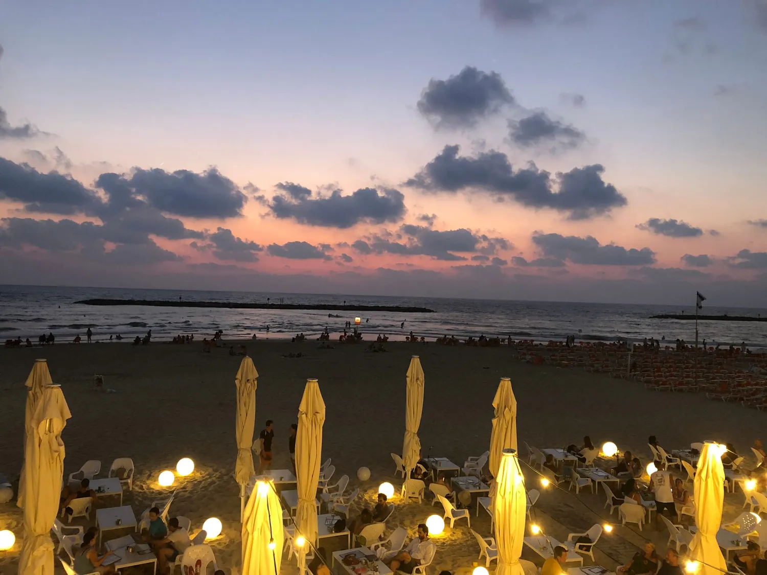 A beach at dusk with umbrellas lit along the sand, a crowd of people out on it and cloud across the sky over the sea.