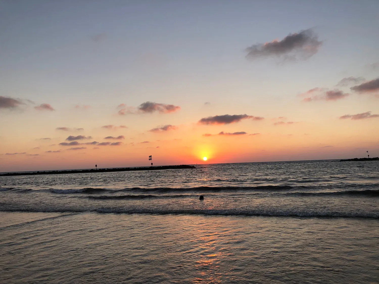 The sun setting between two breakwaters, its light running across the water toward the shore and long lines of swell coming in.