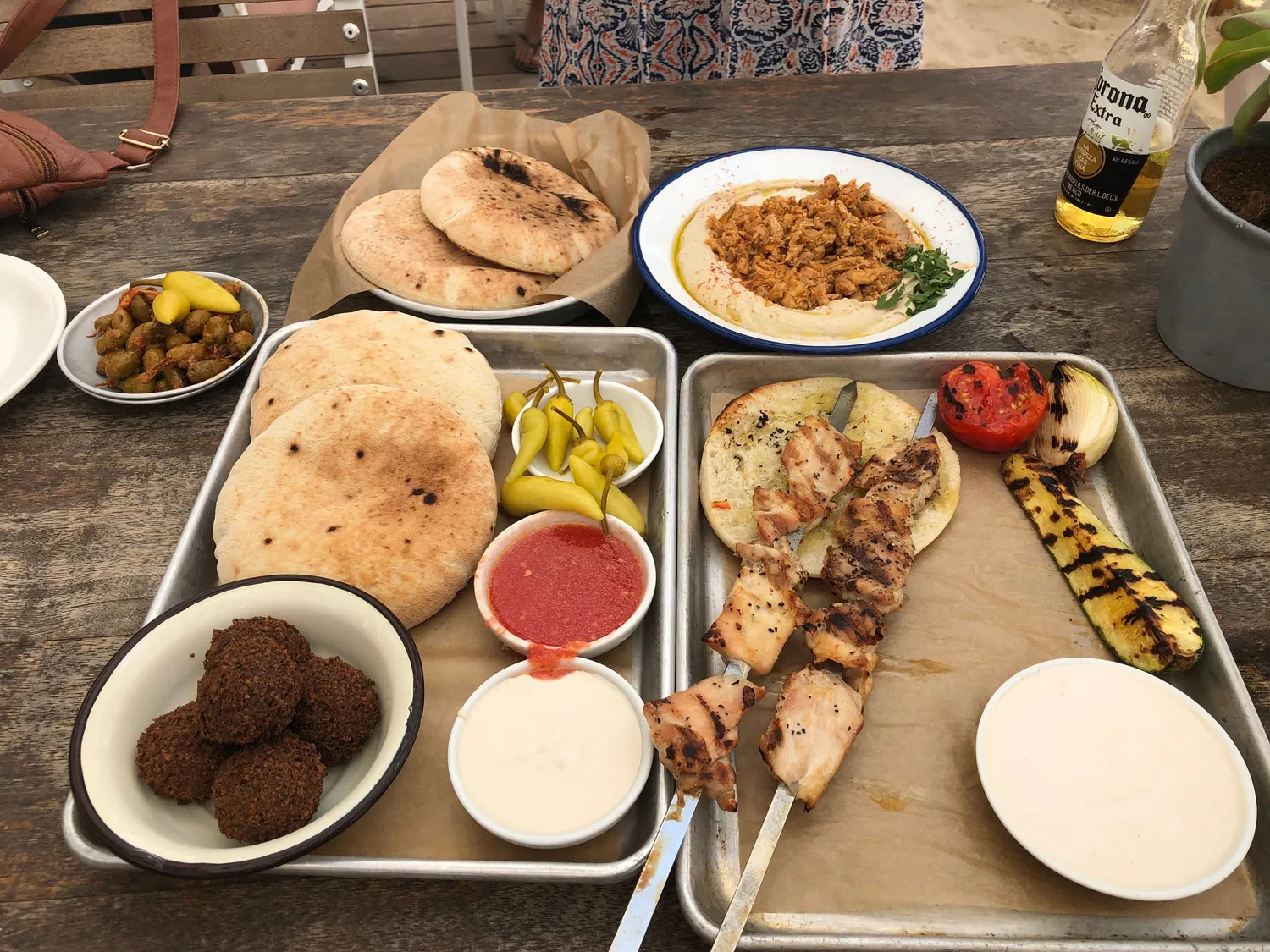 A table laid with flatbreads, bowls of dips and falafel, trays of skewers and grilled vegetables and a tall iced drink, a bottle standing at the end of it.