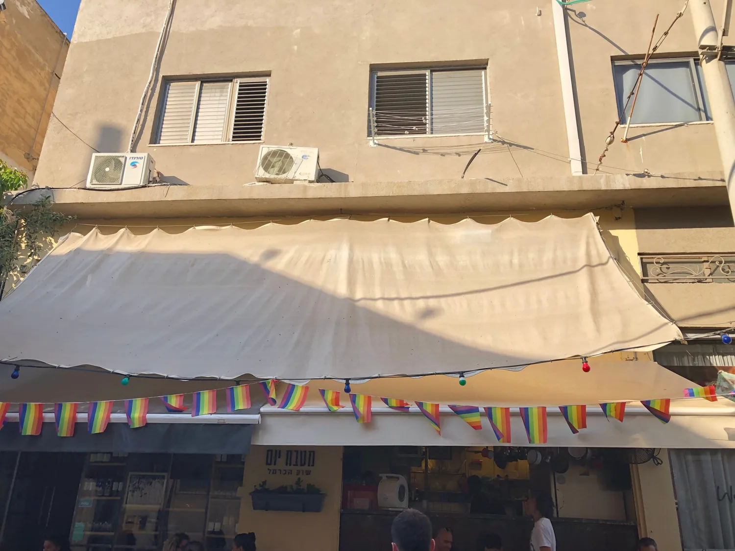 A wide awning stretched out over a pavement, a line of small pennants strung across the shopfront below it, air conditioners on the wall above and lettering over the window.