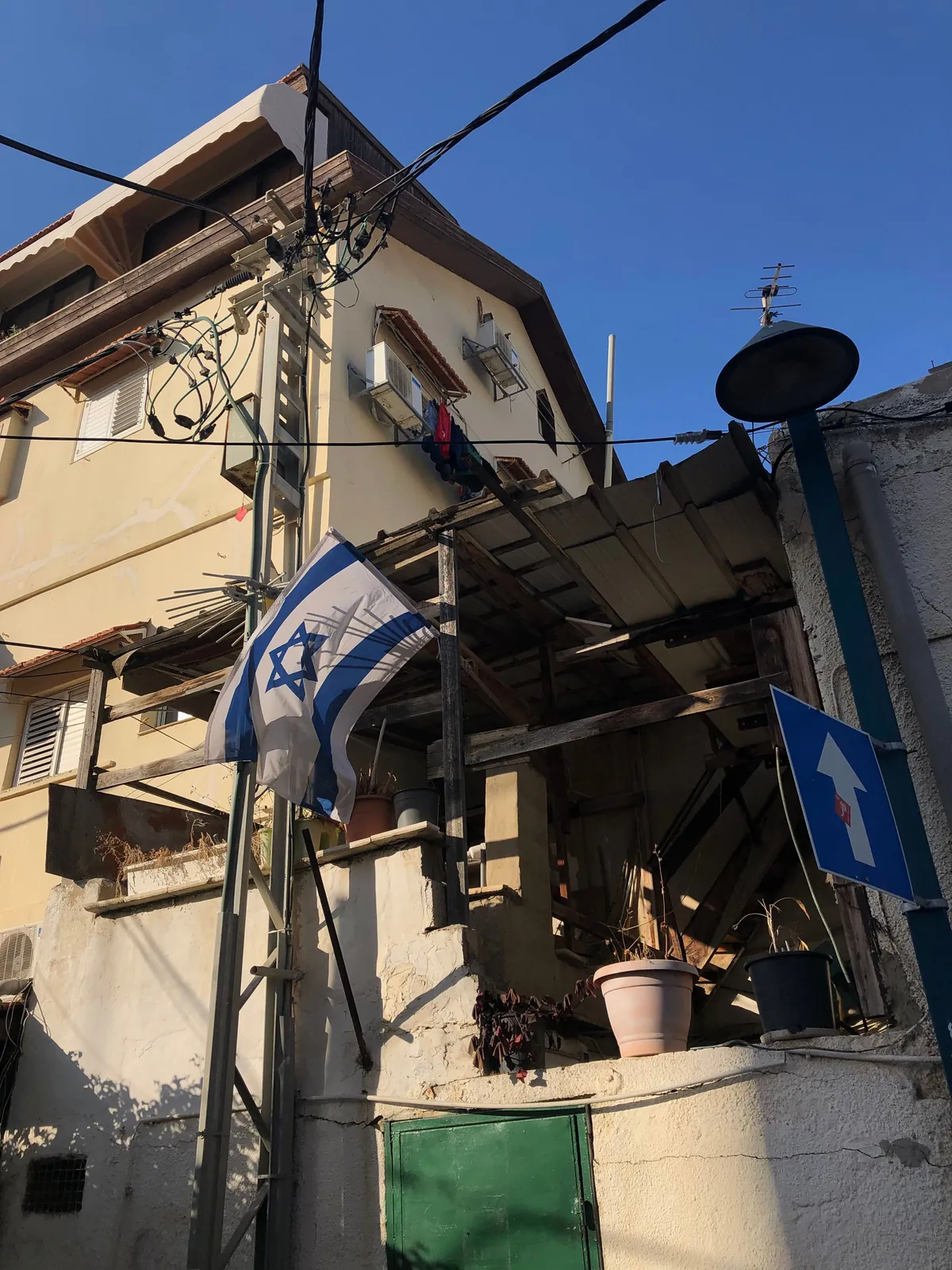 The corner of an old house with a flag on a pole above it, a timber frame set against the wall, wires running across a clear sky and potted plants on the step below.
