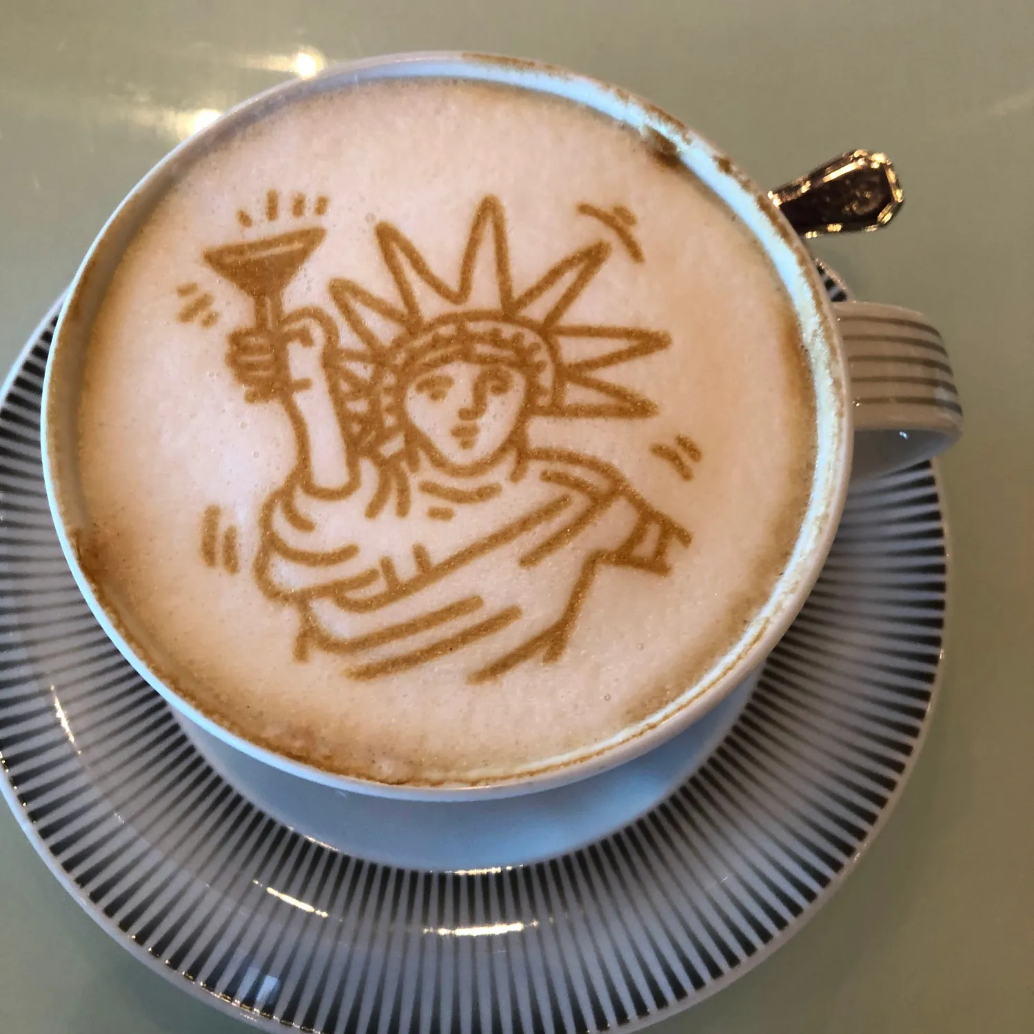 A cup of coffee on a striped saucer with a figure holding something up drawn in the foam, and a spoon resting on the rim.