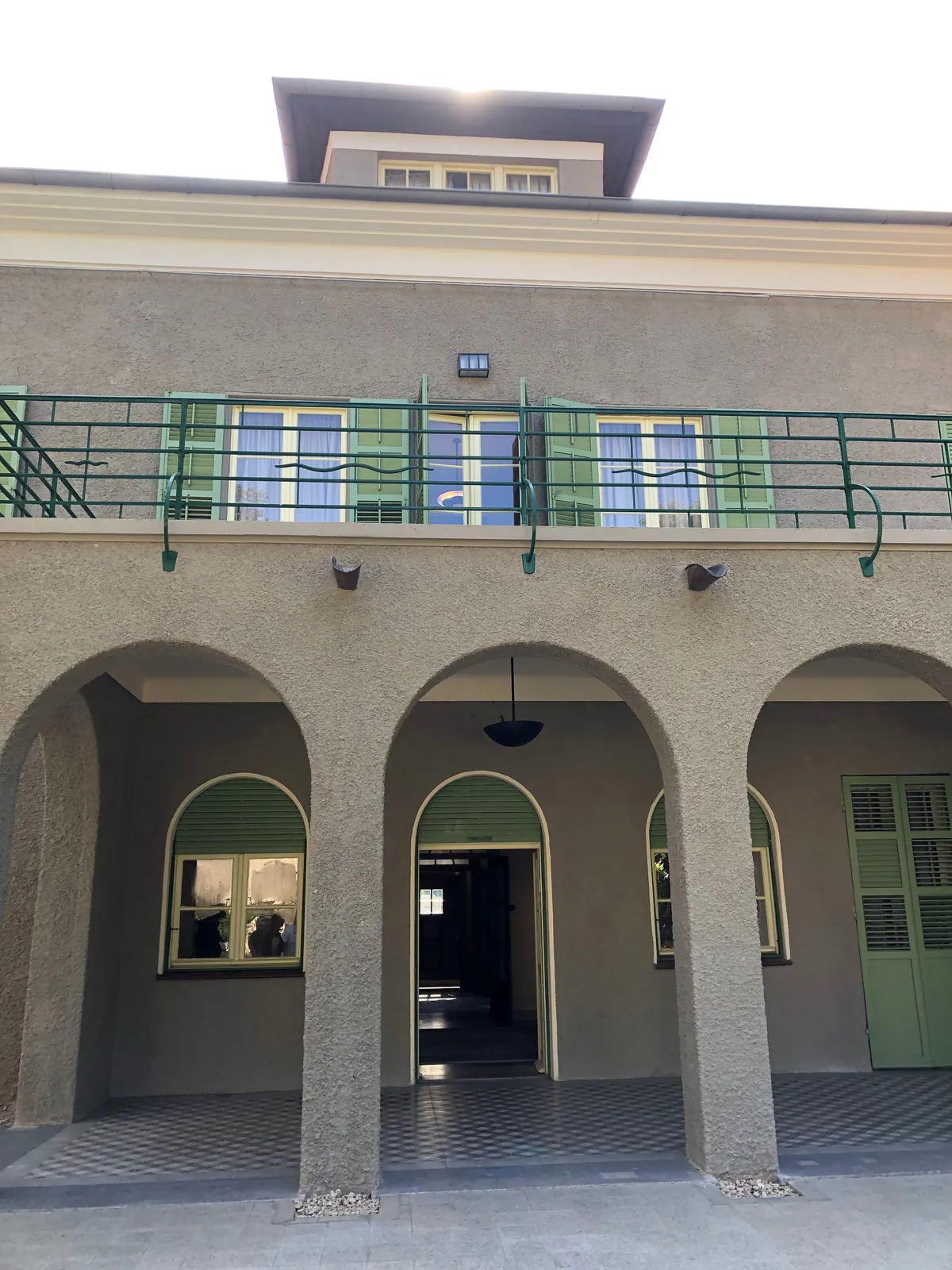 A stone building with three arches along its front, a balcony with a green rail above them, a green door at the centre of the colonnade and a tiled floor running in front.
