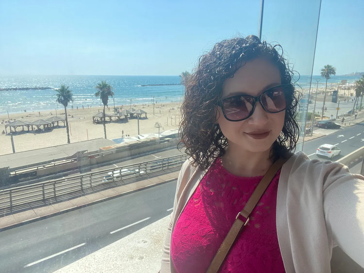 A selfie of a woman with dark curly hair and dark glasses in front of a glass wall, with a sandy beach, a promenade road and the sea behind her.