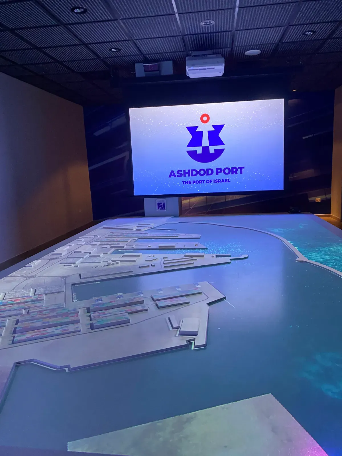 A dark room with a large scale model of a port spread across a table and a big screen behind it showing the port's own emblem and name.