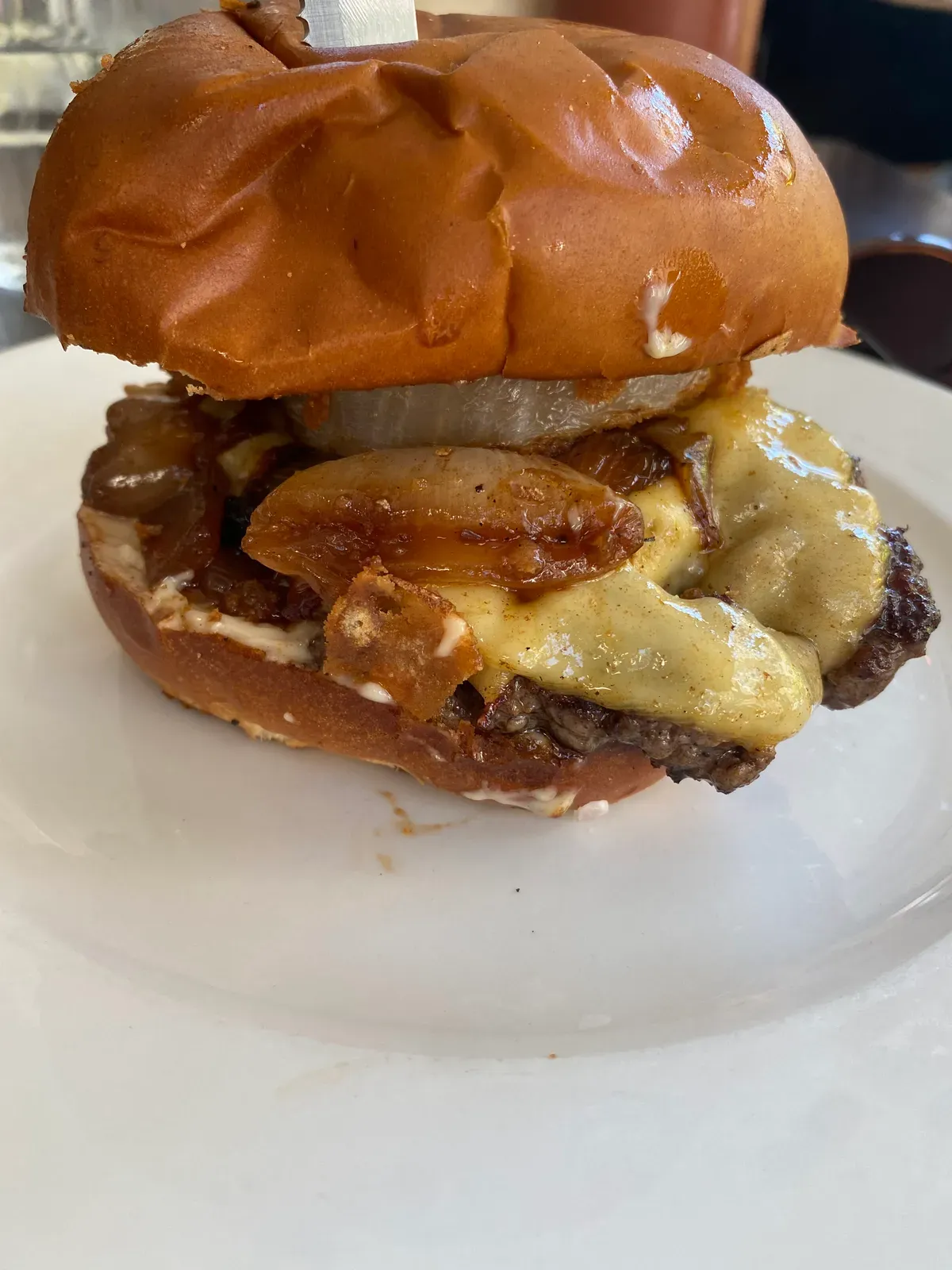 A burger on a white plate on a white table, close up, with cheese and onions coming out over the side.