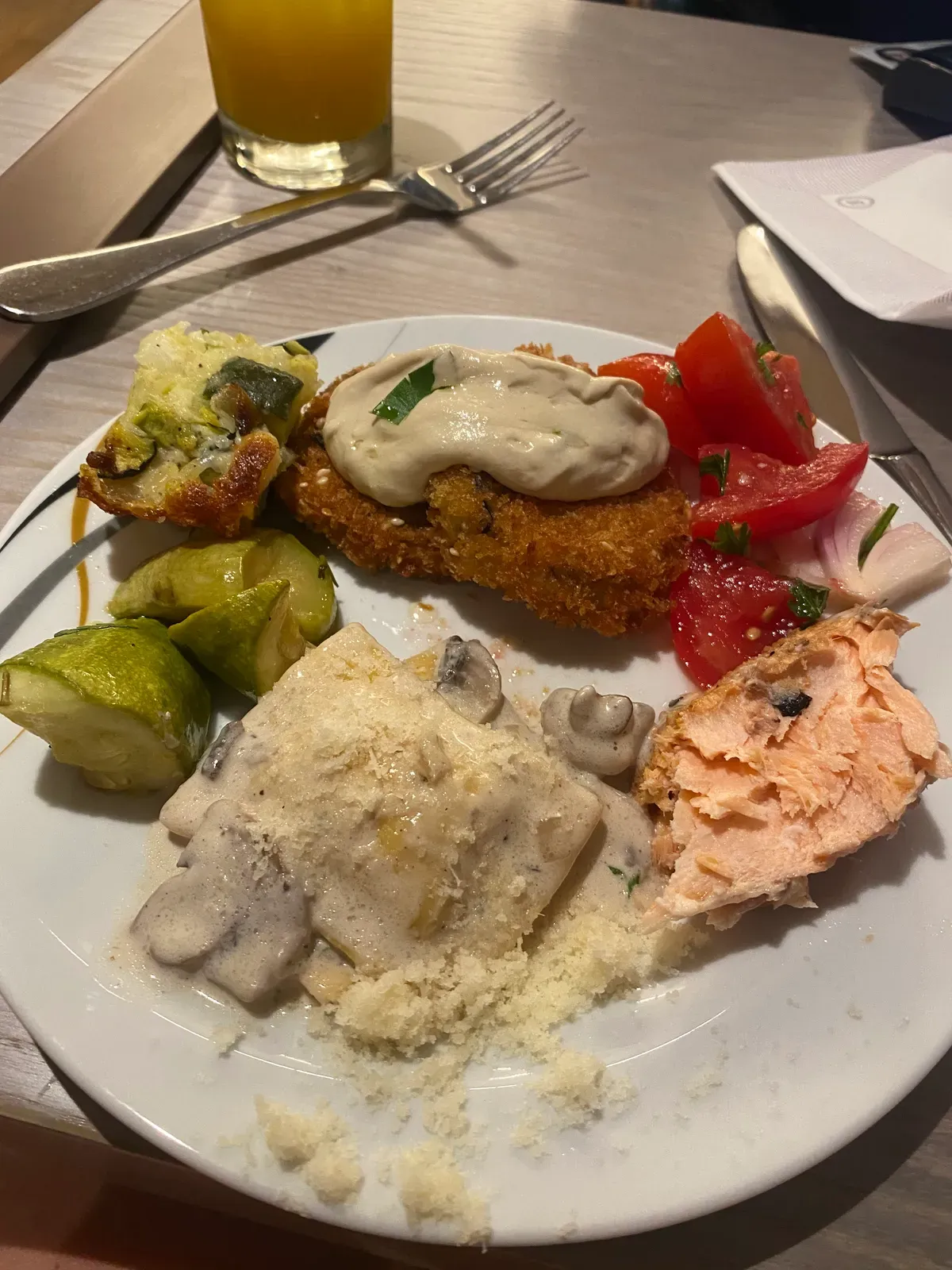 A plate with a breaded cutlet, roasted sprouts, tomatoes, salmon and mashed potato on it, a glass of orange juice behind and cutlery on the table.