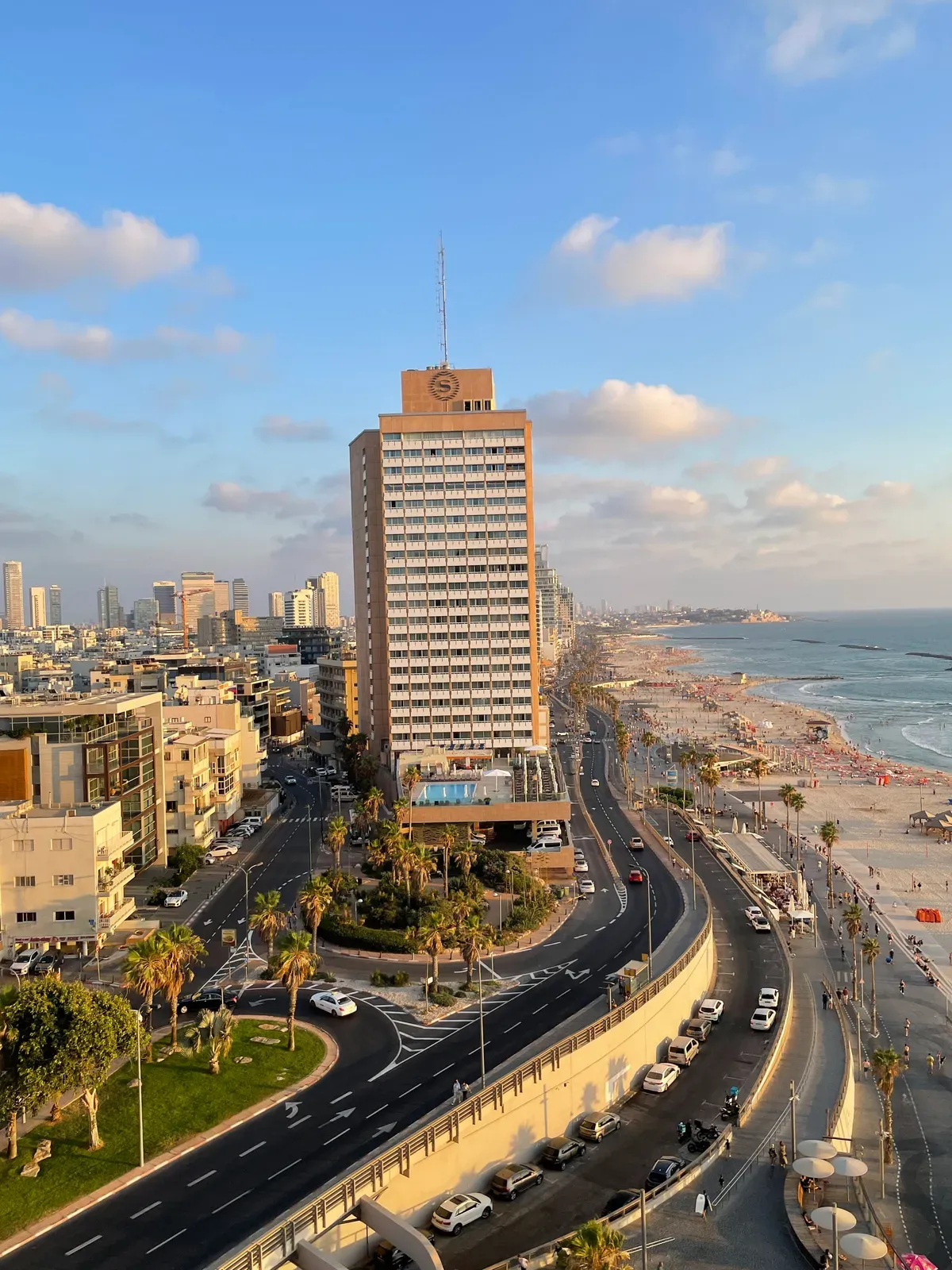 A view down over a seafront in golden evening light, a tall tower with a rooftop sign in the middle, roads and pavements below, and the beach and the sea curving away on the right.