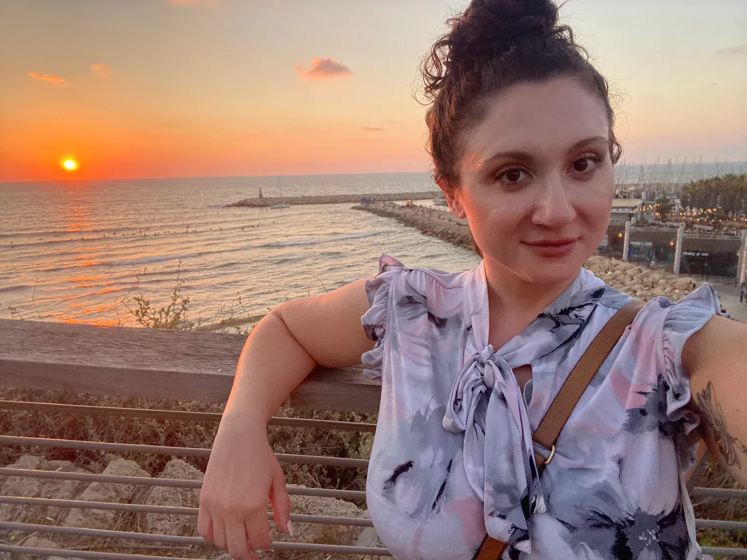 A selfie of a woman leaning on a wooden railing at sunset, the sun low over the water behind her and a breakwater with a small structure at the end of it.