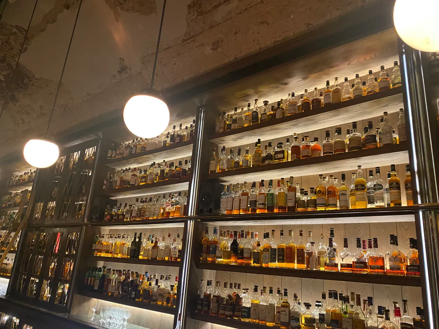 A bar's back wall at night, shelves and shelves of bottles lit from below, round pendant lights above and a dark counter in front.