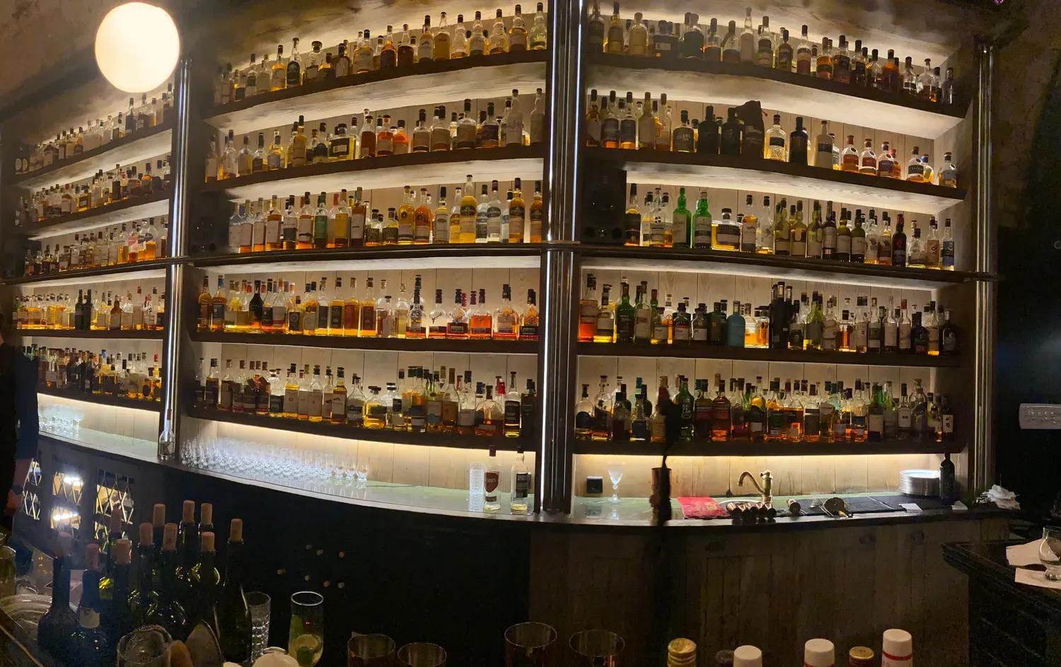 A wide view along a bar's lit shelves, rows of bottles on every level above a counter with glasses, taps and bottles set out along it.