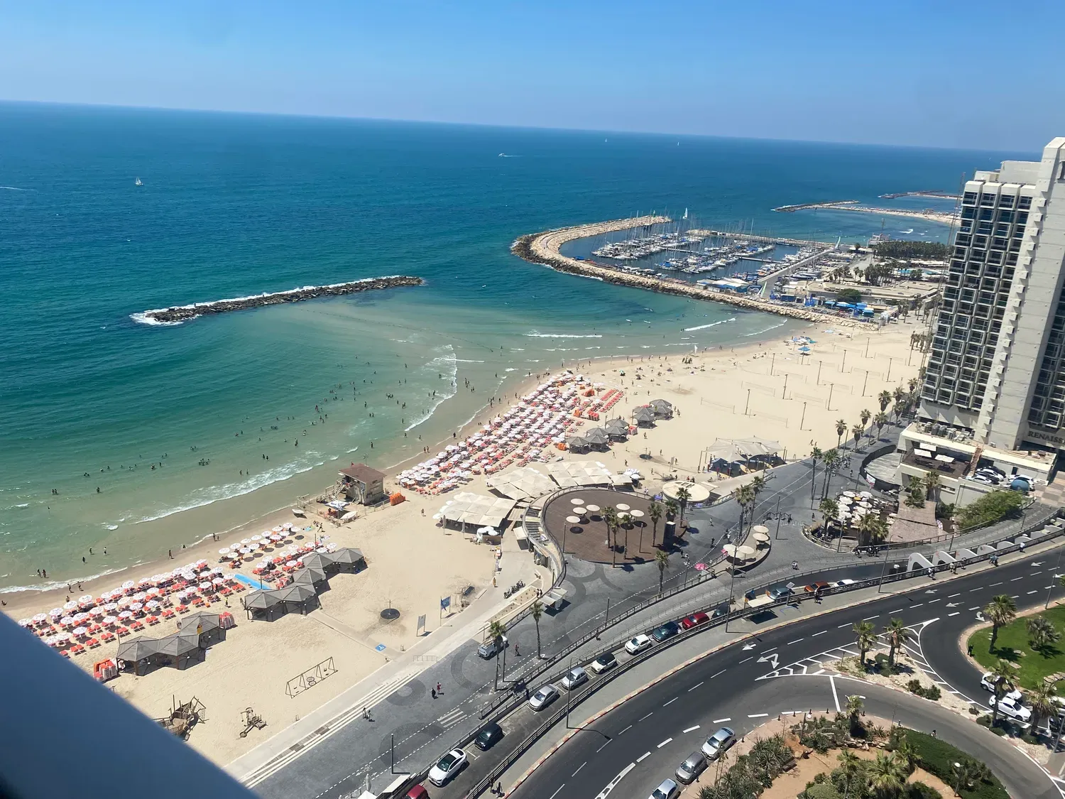 A beach from above on a bright afternoon, rows of sun umbrellas on the sand, a marina full of boats behind a breakwater, and a road along the seafront.