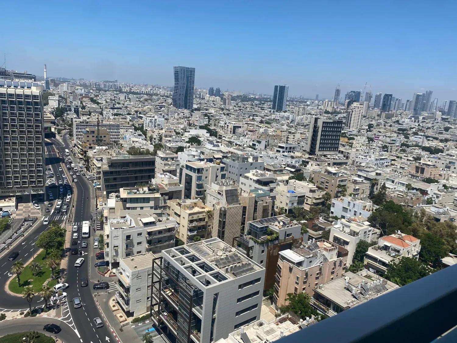 A city from above in daylight, rooftops and streets packed together, traffic on the road below and towers out to the horizon.